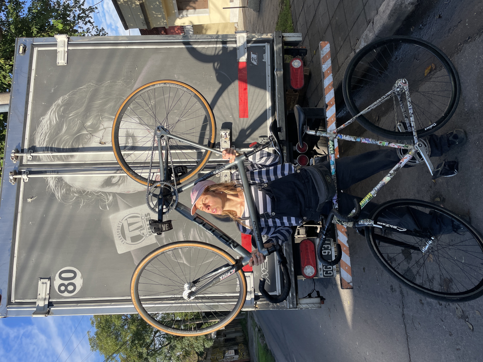 A young person, likely a woman, is captured in a vertical, brightly lit outdoor scene in Florida Oeste, Argentina, posing with two bicycles against the rear of a large gray truck. The subject wears a striped long-sleeve shirt, dark overalls, and a light pink baseball cap, with their eyes squinted and mouth pouted, suggesting a playful or relaxed expression in the sunshine.
One bicycle, with a sleek gray frame, is held upright by the person, its front wheel with a light-colored sidewall prominently displayed. Text visible on this bike includes "DOLAN", "ALPINA", and "PRE CURSA". The other bicycle, positioned closer to the ground, has a frame heavily adorned with an array of colorful stickers, featuring text such as "VAMP", "Dialcez", and "BkZam". Both bicycles have tires marked "DOWNHILL ALOY".
The rear of the gray truck serves as a backdrop, featuring a large, faded graphic of a person's face with wavy hair. A red stripe runs across the truck's lower panel, and a circular "80" speed limit sign is visible on the left side. The truck's license plate, located on the right, reads "GG 93". Additional text "TRANSPORTE" is visible on the truck, and "lubri" appears to be on a component of one of the bikes. The scene is bathed in bright daylight, casting distinct shadows. Trees and a clear blue sky are visible in the background, suggesting good weather.