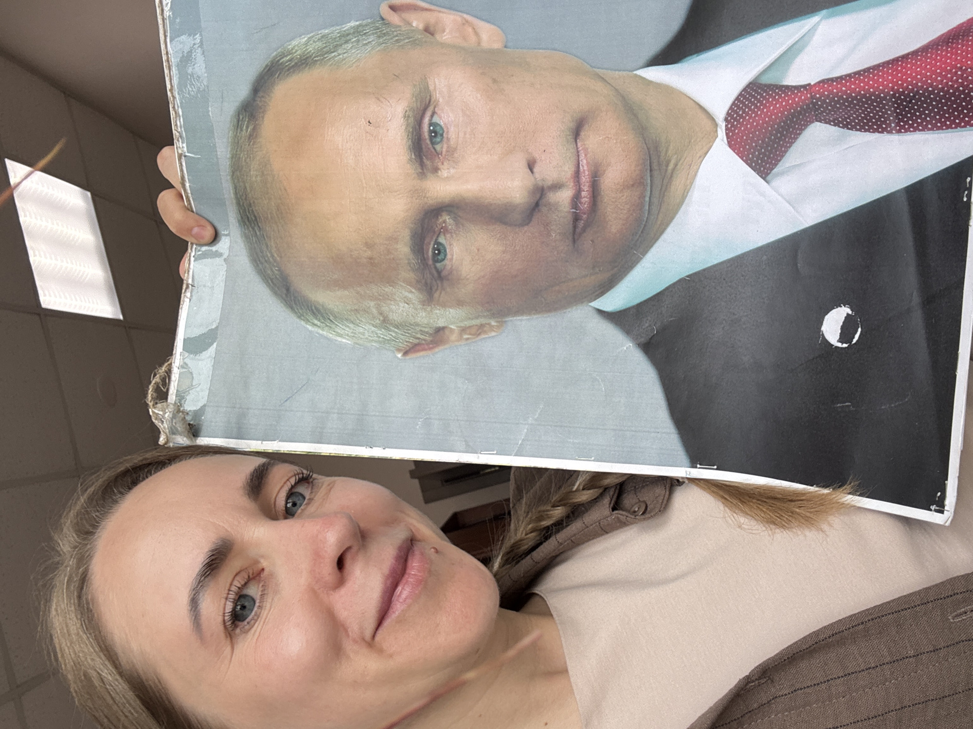 A woman is holding a damaged poster of Vladimir Putin. The poster shows a portrait of Putin wearing a black suit, white shirt, and red tie. The woman has blue eyes and blonde hair partially braided. She is wearing a beige shirt and a brown striped jacket. The background appears to be an indoor setting with white walls and recessed lighting. There is no visible evidence to confirm the location in Chaikovsky, Russia.
