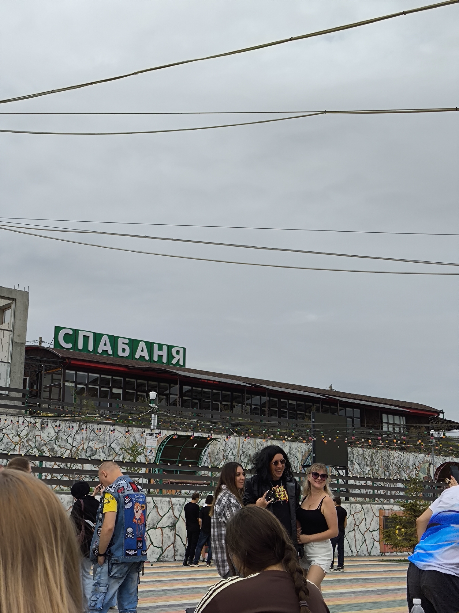 The image depicts an outdoor scene in Borgustanskaya, Russia, under an overcast sky. In the background, a building with a long, dark roof is visible, featuring a green illuminated sign that reads "СПАБАЯНЯ". Several people are gathered in front of the building. Among them, a person with long dark hair and sunglasses, wearing a black leather jacket, is interacting with two women, one with blonde hair and sunglasses, wearing a black tank top and white shorts, and another woman in a plaid shirt. To the left, a man wearing a denim vest covered in patches and a yellow t-shirt is facing away from the camera. The environment includes a decorative stone wall with lights strung across it, and trees or bushes behind a fence. There are also overhead wires visible against the sky.