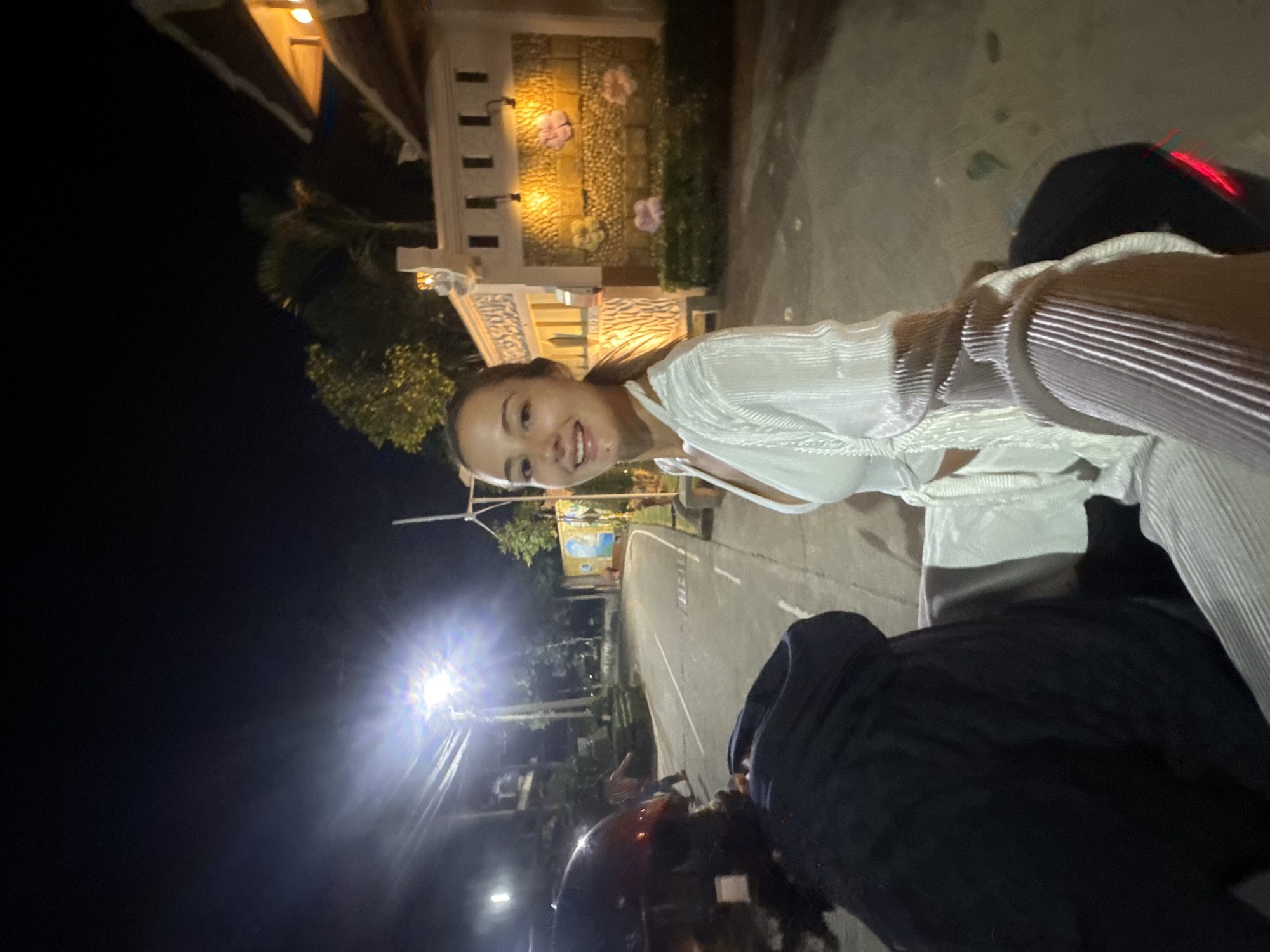 The image shows a woman in a white outfit sitting on a motorcycle in Pattaya City, Thailand at night. The background features a building with decorative lighting and foliage, as well as street lights illuminating the road.