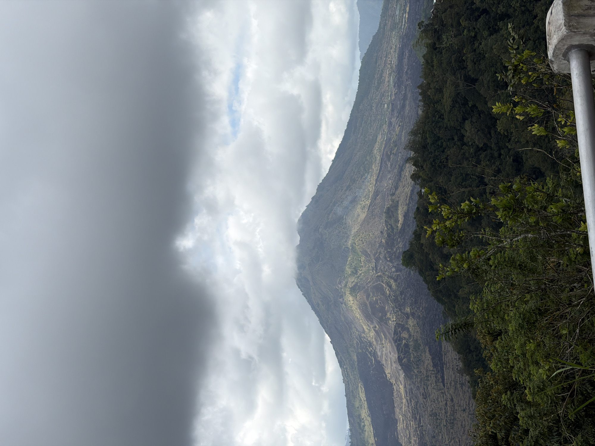 The image shows a panoramic view of Mount Batur in Kintamani, Indonesia. The volcano is large and imposing, with a rugged, dark landscape surrounding its base. Sparse vegetation, mostly green shrubs and trees, grows on the lower slopes. The sky is overcast with heavy gray clouds, obscuring the sun and casting a somber light on the scene. In the foreground, a concrete barrier with a metal railing is visible, suggesting this is a viewpoint or overlook. The overall impression is of a dramatic and wild natural landscape under a moody sky.