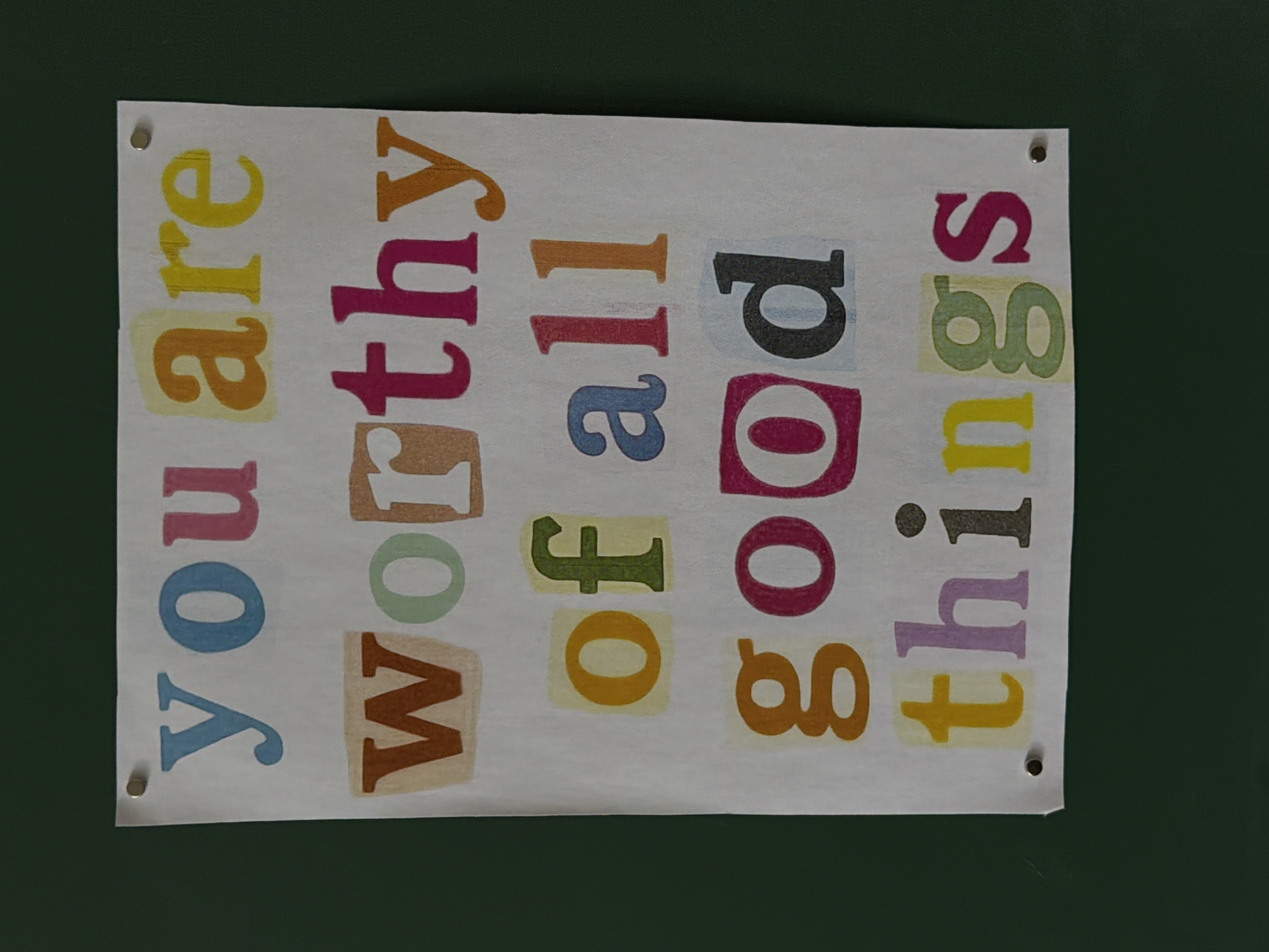 A white rectangular sign with colorful letters spelling out "You are worthy of all good things" is affixed to a dark green surface. The sign is held in place by tacks or pins visible at the top left and right corners, and bottom right corner. The background is a solid dark green with no other visible elements. The location is not specified.