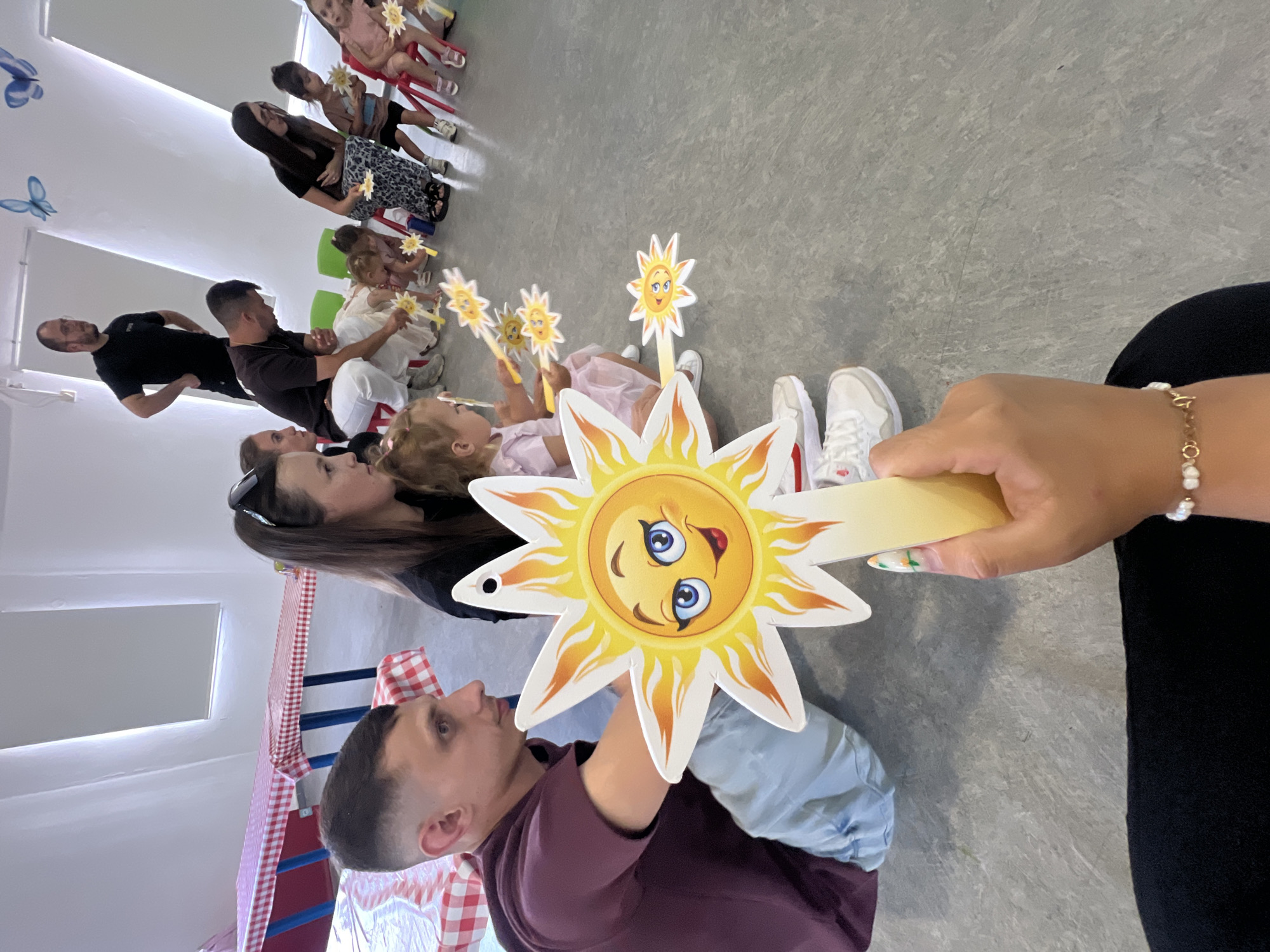 This image depicts a group of people, a mix of adults and children, gathered indoors in what appears to be a classroom or event space. The subjects are arranged in a semi-circle on the floor, with some seated on small chairs. They are holding and looking at yellow cutouts shaped like the sun with smiling faces on sticks. One large sun cutout is held prominently in the foreground by an adult whose hand and arm are visible. The children are dressed in casual clothing, and some appear to be wearing costumes or themed outfits. The adults are also in casual attire.
The scene is well-lit, suggesting it is daytime, and there are no clear indications of the weather outside. The room has a light-colored floor and white walls. In the background, there are some decorative blue butterflies affixed to the wall and a table covered with a red and white checkered tablecloth. The overall atmosphere seems to be one of a playful gathering, possibly a children's party or a school activity. The people in the image appear engaged and some are smiling, contributing to a positive and cheerful mood. The location context is Kiryat Bialik, Israel, though there are no specific visual cues in the image to confirm this. No visible text is present in the image.