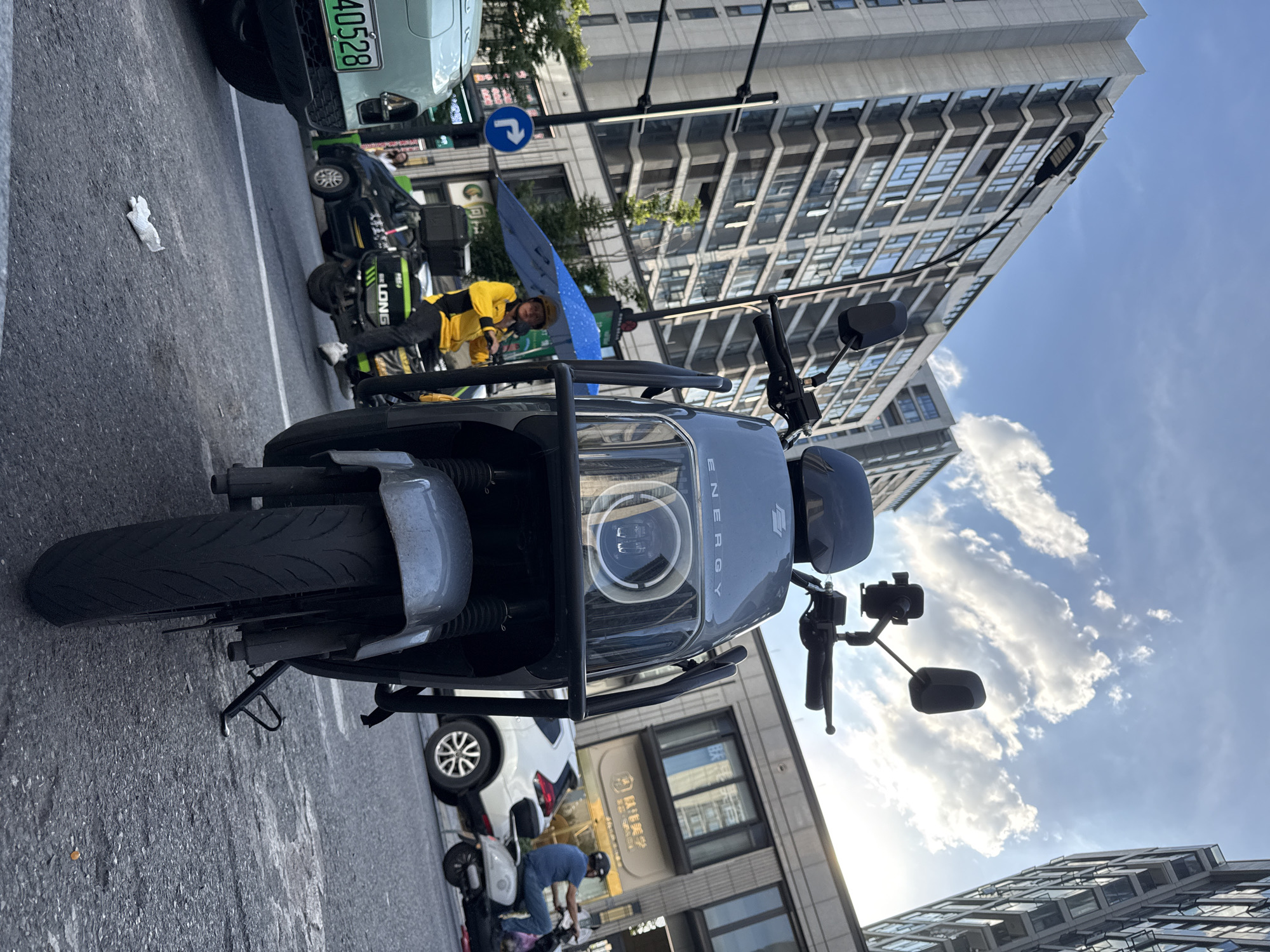 The image captures a street scene in Yiwu, China, under a partly cloudy sky. In the foreground, the front of a gray electric scooter is prominently displayed, with its headlights illuminated. Behind the scooter, a delivery person in a bright yellow jacket and helmet is holding a blue umbrella, seated on a black electric scooter. This scooter is equipped with a large black storage box on the back labeled "LONG." To the left of the scene, a portion of a light green car with a license plate reading "40528" is visible. In the background, tall, modern buildings with numerous windows line the street. A white car is parked on the right side of the road, and another individual on a scooter can be seen in the distance. The asphalt street shows signs of wear and tear, and a crumpled white paper object is lying on the ground in the mid-ground. A blue directional sign with arrows is mounted on a pole. The overall impression is a snapshot of daily urban life in Yiwu, with a focus on transportation and commercial activity.