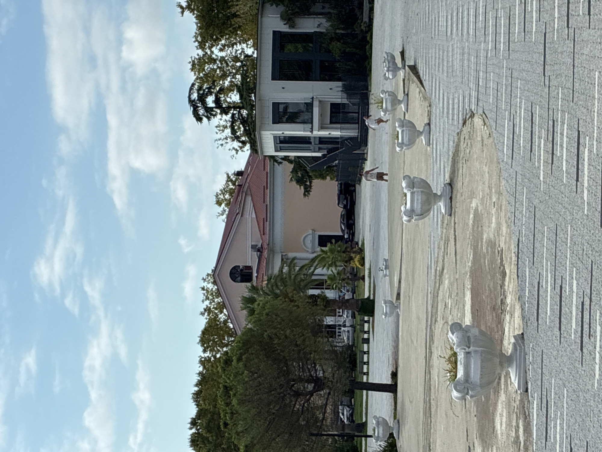The image depicts an outdoor scene in Sochi, Russia, featuring a large pale yellow building with a red roof and a modern white building with dark-framed windows. Several large white planters are visible on a paved area in the foreground. Two children, one in a white dress and the other in shorts and a t-shirt, are running in the middle ground. Trees, including a pine and palm trees, are scattered throughout the scene. Cars are parked in the background. The sky is blue with scattered white clouds.