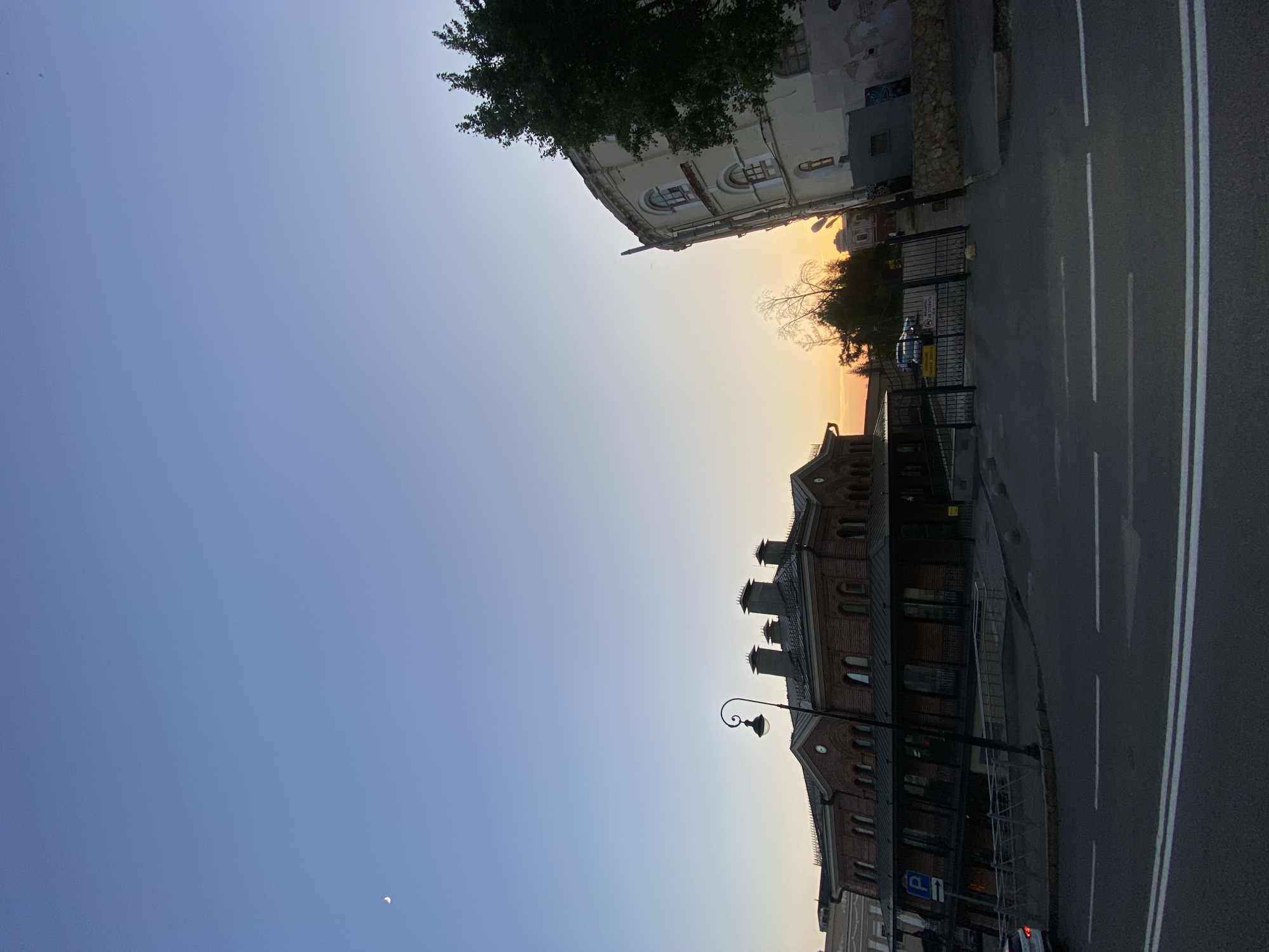 The image shows a street scene in Kazan, Russia at dusk. A multi-story brick building with distinctive chimneys lines one side of the street. A lamppost stands in front of the building. Across the street is a lighter-colored building with arched windows. A road with double white lines runs through the scene. The sky transitions from blue to yellow near the horizon. A crescent moon is visible in the blue part of the sky. A fence can be observed between the old brick building and the road. There is a car on the road, and some parked vehicles.