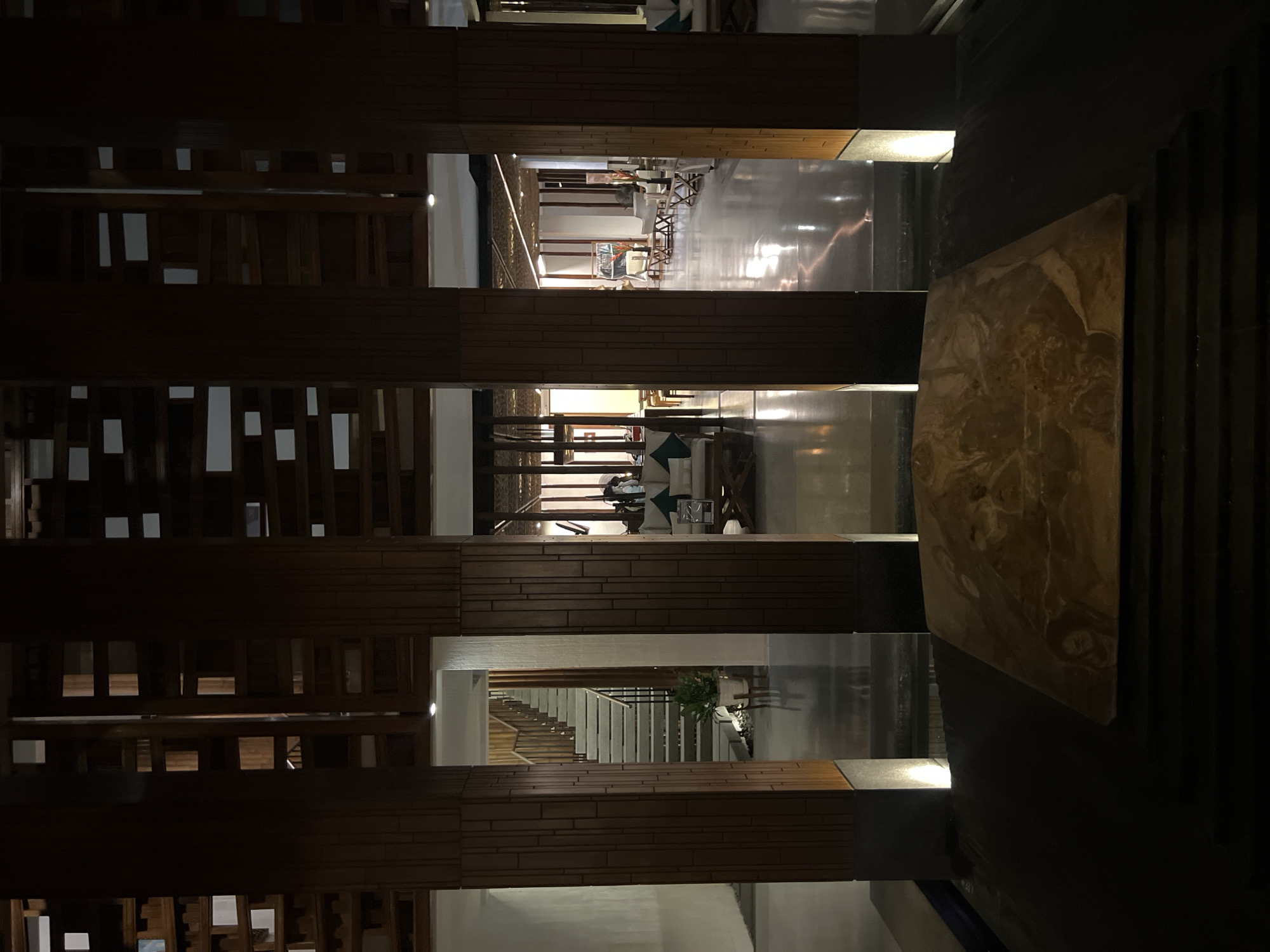 The image depicts an interior architectural space, likely a hotel lobby or a similar establishment, at night. Dominating the foreground are four large wooden pillars, creating a sense of depth and separation within the space. Between these pillars, the scene opens up to reveal more of the interior.
To the left, a staircase with wooden railings ascends, illuminated by overhead lighting. A potted plant sits at the base of the stairs.
In the center and to the right, the view extends into a lounge area. Sofas and seating arrangements are visible, with coffee tables and decorative elements. The flooring is highly reflective, creating mirrored images of the lighting and architecture. The ceiling features an intricate, textured design with embedded lights, contributing to the ambient illumination. The overall atmosphere is dimly lit, with warm light sources highlighting different areas of the space. The scene is devoid of any visible people. The material of the large square object in the immediate foreground appears to be stone or marble with a polished surface.