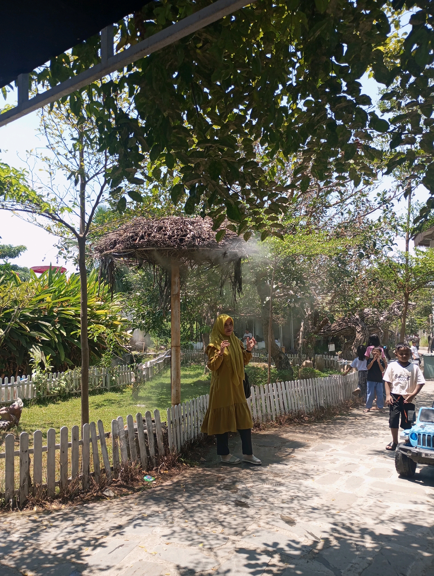 In the city of Ponggok, Indonesia, a sunny outdoor scene unfolds. The foreground features a paved pathway with dappled sunlight filtering through overhead foliage. To the left, a white picket fence lines a grassy area with lush green bushes and trees. A woman in a yellow hijab and a long-sleeved dress stands near the fence, looking to her right, with a black bag slung over her shoulder. In the mid-ground, more greenery and a straw-covered canopy supported by poles are visible. A fine mist is being sprayed from the canopy, creating a hazy effect. Several people, including children, are present in the background, some walking and others standing. A small blue toy jeep is parked on the pathway to the right. The overall environment suggests a recreational area, possibly a park or tourist attraction, with a focus on natural elements and a relaxed atmosphere.