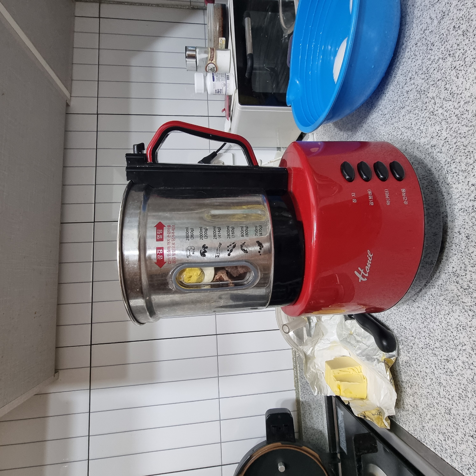The image depicts a kitchen scene, likely in a residential setting. A red soup maker, branded "Hance," is the central object. It has a stainless steel inner pot with measurement markings visible through a clear window, containing what appears to be food ingredients. The soup maker sits on a speckled countertop. To the left of the soup maker, a block of butter is unwrapped, resting on its packaging. Behind the soup maker, a white microwave oven is visible, with a white pill bottle, a salt shaker with a rosemary label, and a small jar of dark sauce on top of it. To the right of the soup maker is a large blue plastic bowl, partially filled with water and some white items. The background consists of white tiled walls with gray grout lines, and a light-colored ceiling. The overall impression is of a domestic kitchen ready for meal preparation. There is no indication of the specific location within Dunnae-myeon, South Korea.