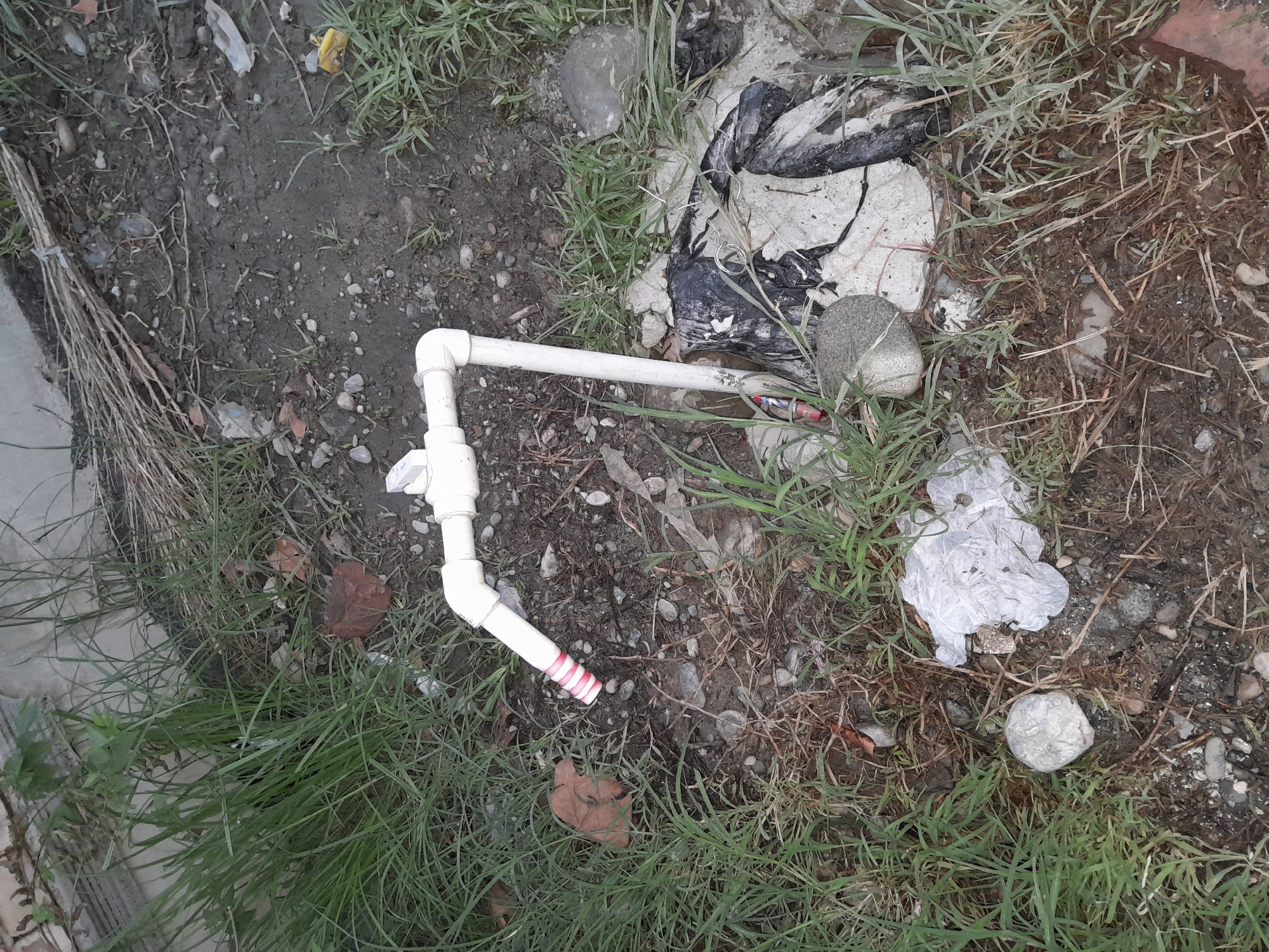 The image shows a close-up view of a white plastic pipe with a faucet attached, protruding from the ground. The pipe is bent at a right angle and appears to be part of an outdoor water supply. It is surrounded by dry grass, dirt, rocks, and some debris, including pieces of plastic and a red brick. The scene is outdoors and appears to be in a somewhat neglected area, possibly a yard or an unpaved public space. There are no people visible in the image. The overall impression is one of an ordinary, unexceptional outdoor setting.