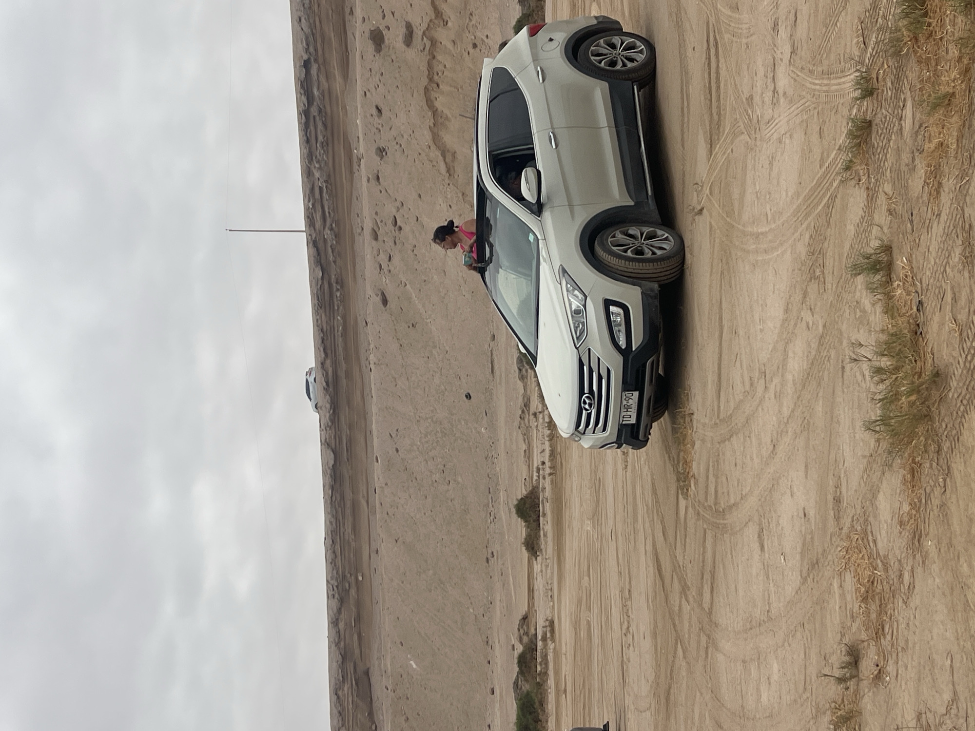 A white Hyundai Santa Fe is parked in a sandy, off-road environment with visible tire tracks, likely near a coastal desert setting. A woman with dark hair pulled back and wearing a pink top is leaning out of the driver's side window. The terrain appears to be dry and arid, with sparse vegetation. In the background, there is a barren hillside or dune and utility pole. The license plate is visible, reading "TD-HR-90". The location is likely in or near the Atacama Desert surrounding Arica, Chile, given the arid landscape.