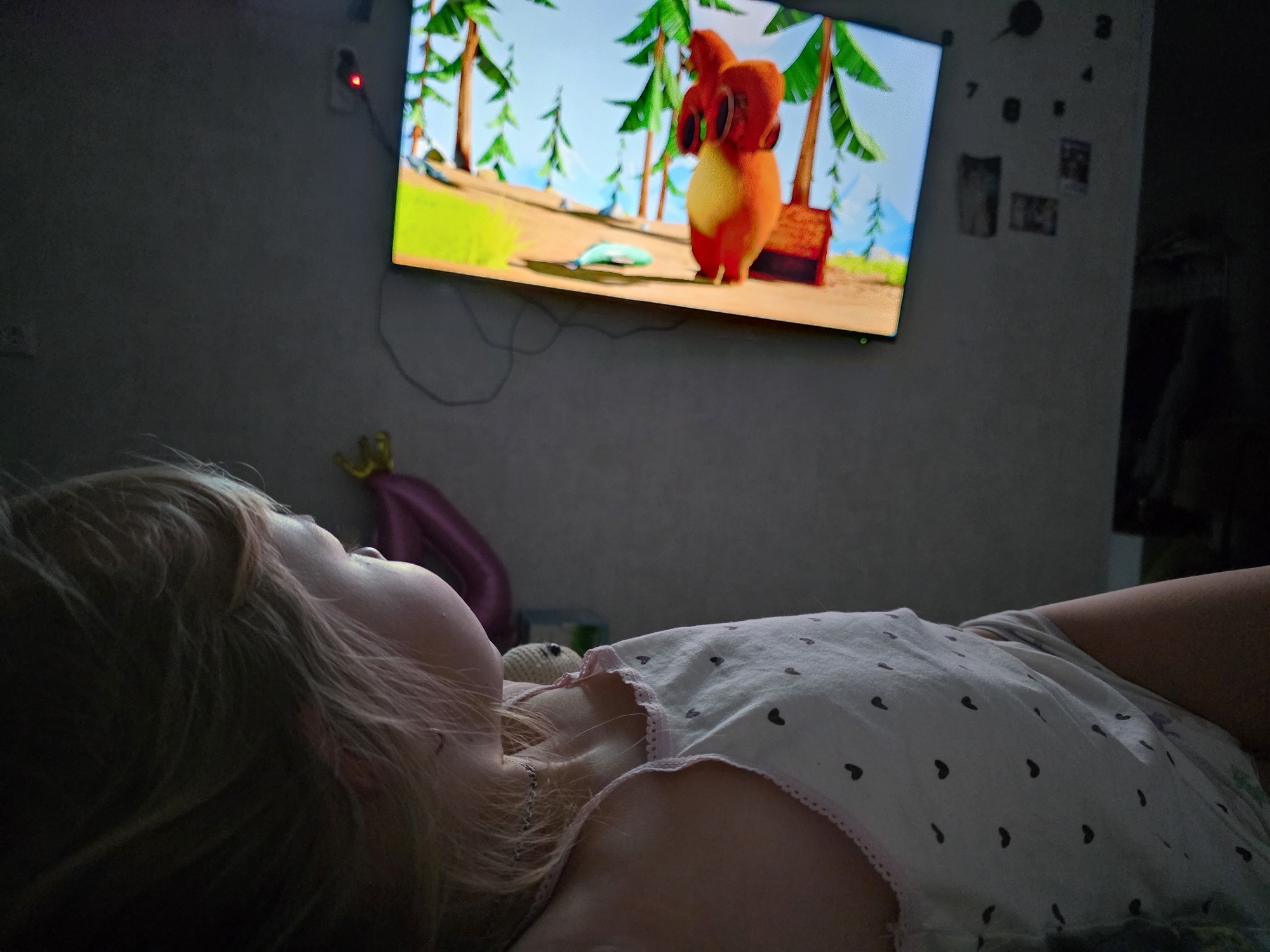 A young, fair-skinned child with blonde hair lies on a bed. The child's head is turned upwards, facing a wall-mounted television displaying an animated cartoon with a red, fuzzy character in a tropical-like scene. The child is wearing a white shirt with small, dark-colored heart patterns and a pink scalloped edge. Visible behind the child is a partially visible purple balloon and other indistinct objects.
On the wall to the right of the television are dark, abstract clock numbers and a few small, indistinct photographs or prints. The overall lighting in the room is dim. The location is the city of Chaikovsky, Russia.
