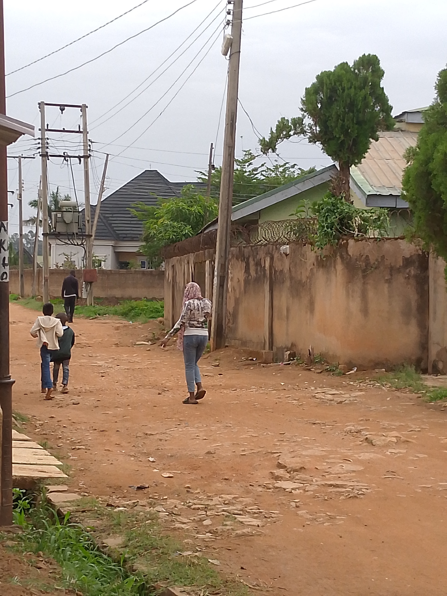 The image depicts an unpaved street in Jos, Nigeria. The ground is reddish-brown and uneven. Three children are walking down the street, followed by an adult. A woman wearing a headscarf and jeans walks in the opposite direction. The street is flanked by buildings and walls, some topped with barbed wire. Power lines and utility poles are visible. Vegetation grows along the edges of the street. The sky is overcast.