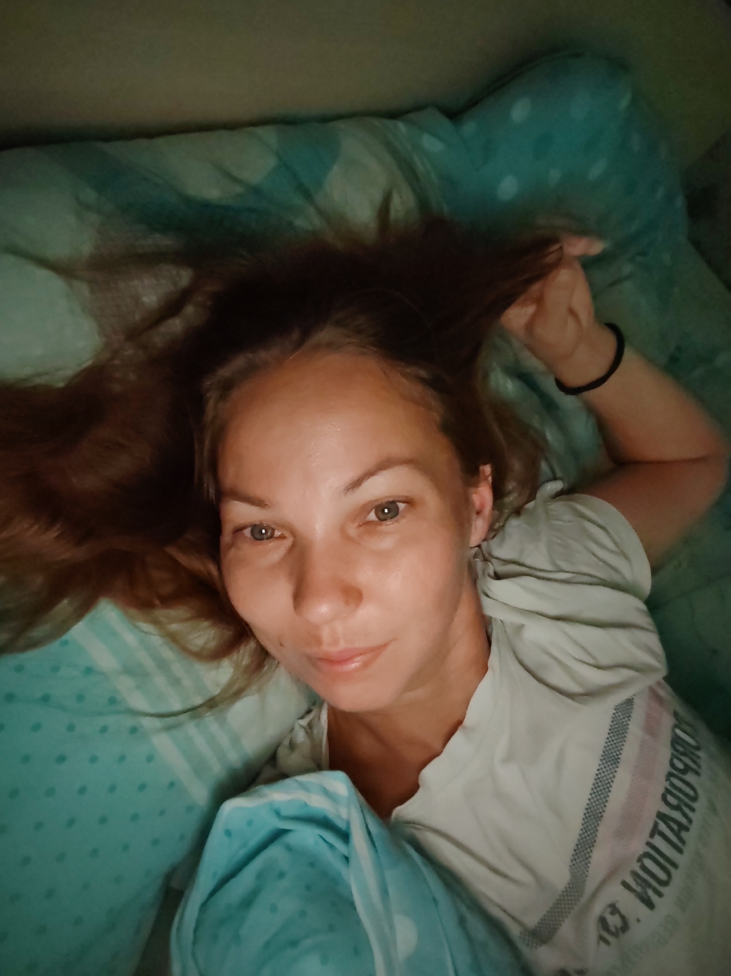 A young woman with long, brown hair lies on her back in bed, looking up at the camera. Her head is slightly tilted to the left, and she is holding a strand of her hair with her right hand. She is wearing a light-colored t-shirt with stripes on the sleeve. The bedding appears to be a light blue color with a polka dot pattern. The overall lighting suggests it may be nighttime or an indoor setting with low light. The image appears to be a selfie taken from a close-up perspective. The background is dark, obscuring any details of the room. There is no visible information within the image to suggest a specific location in Чухлэм, Russia.