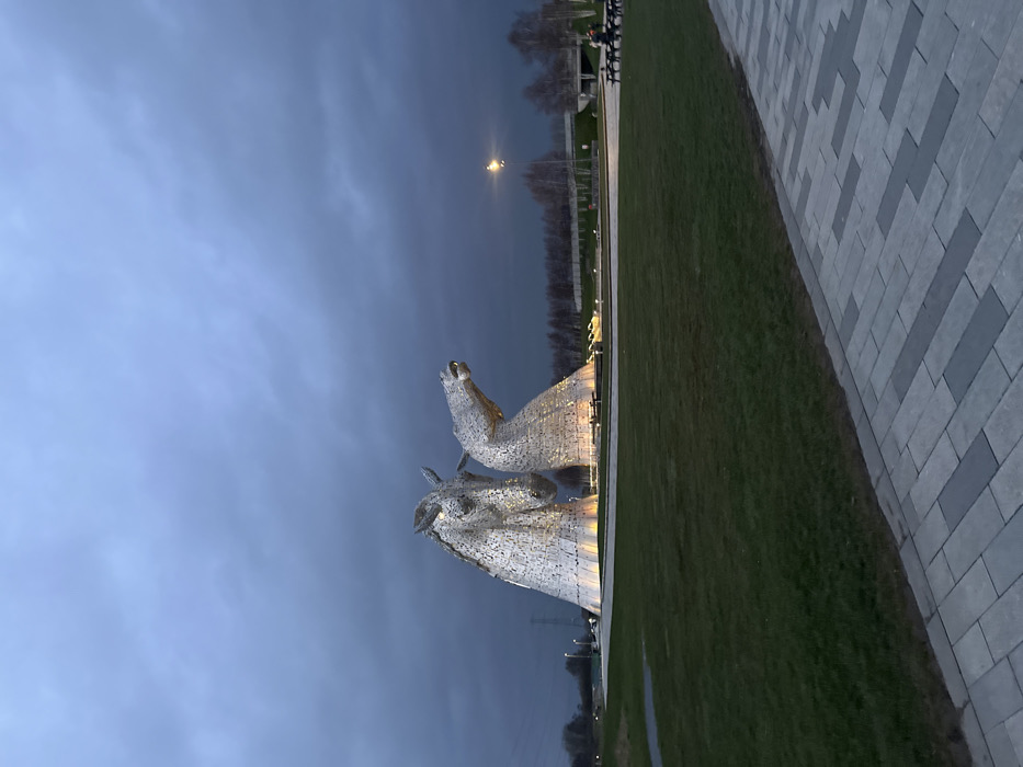 The image displays The Kelpies, two colossal equine head sculptures, illuminated by warm yellow light from their base, against a dark, overcast blue-grey sky. The scene is located in Falkirk, United Kingdom, during evening or twilight hours. The sculptures are composed of numerous metallic panels and stand within a circular water feature or landscaped area. The taller, primary head faces forward and slightly left, while the secondary head is angled downward and to the right. In the foreground, a paved pathway with interlocking light and dark grey rectangular blocks runs alongside a manicured green lawn. In the mid-distance on the right, two individuals are seated at a light-colored wooden picnic table with attached benches. The background includes a line of bare deciduous trees, an elevated road structure, and a bright, distant streetlamp. Faint power lines and industrial structures are visible on the far left horizon.