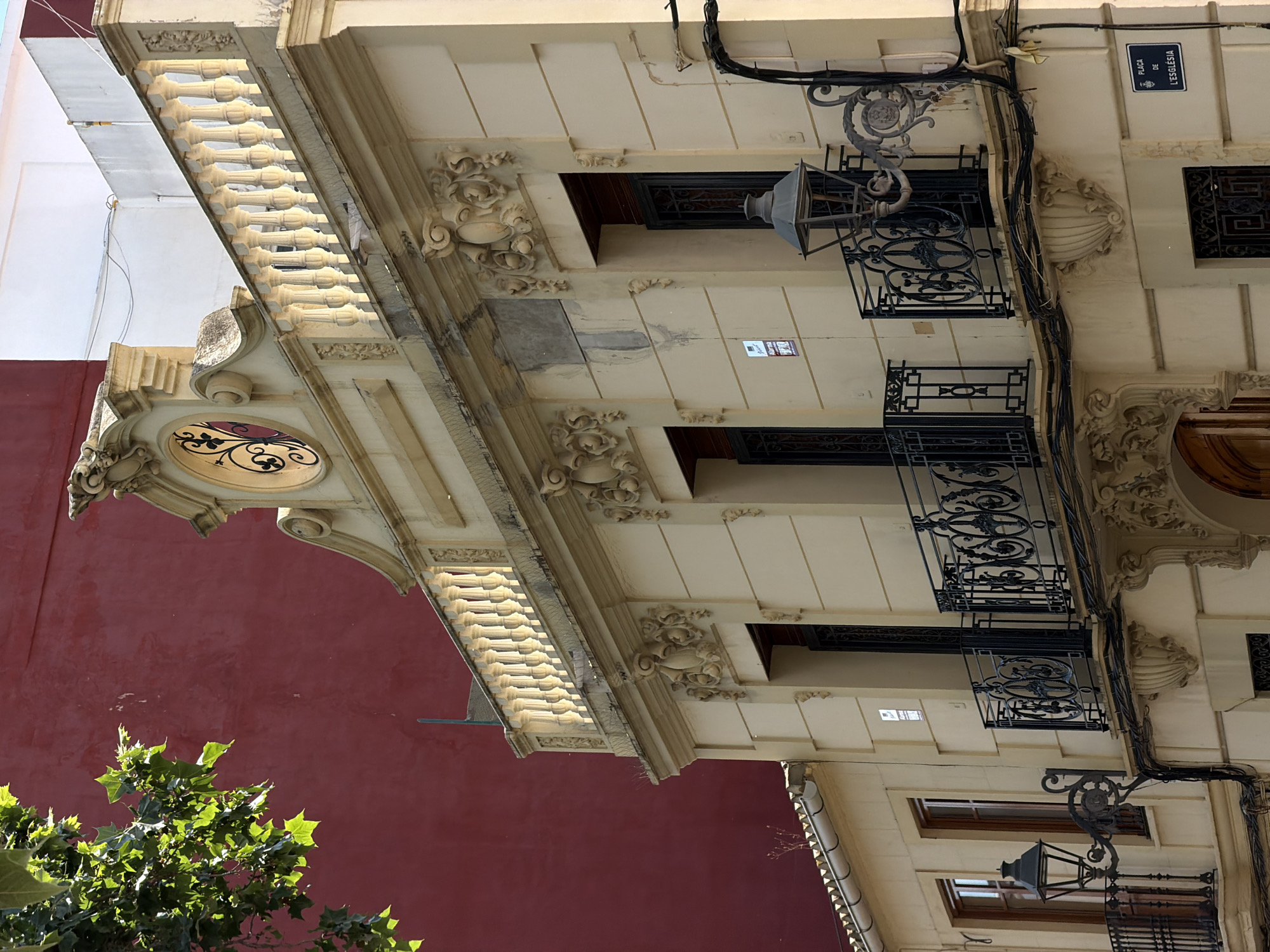 An exterior, eye-level, medium shot captures a beige building facade with ornate architectural details. The building features a central decorative element above a rounded arch, flanked by carved ornamentation. A balcony with black wrought-iron railing extends across the middle of the facade. Another balcony with a similar railing is visible to the left, and a street lamp is attached to the wall near it. The building's facade has multiple windows, some with wooden frames and intricate iron grilles. A blue street sign is affixed to the corner of the building, reading "PLACA DE L'ESGLÈSIA". Above the main balcony, a balustrade with white balusters runs along the roofline. To the left of the building, a large section of a dark red wall is visible, partially obscured by the leafy branches of a tree in the foreground. The sky is a bright, clear blue.