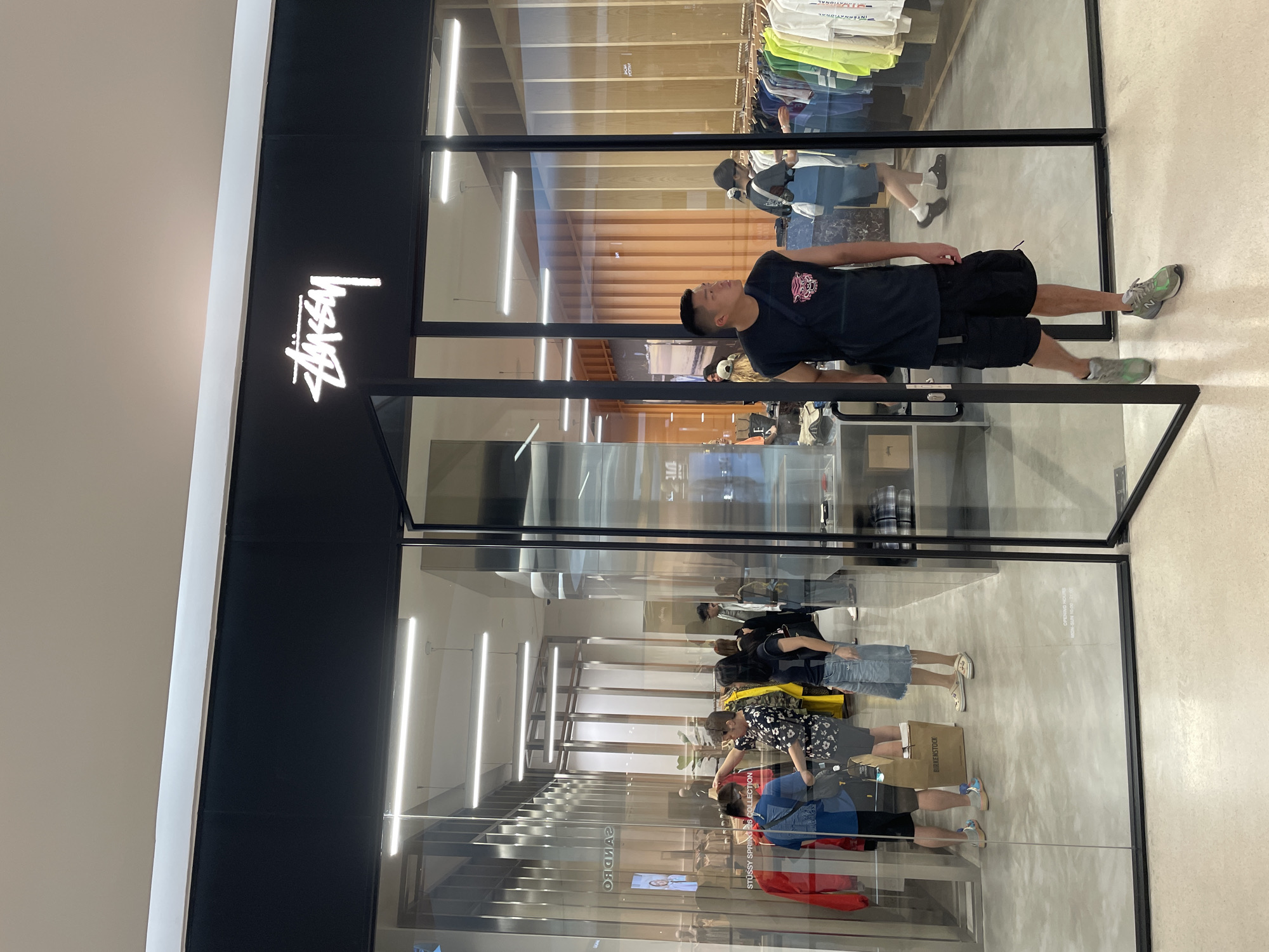 The image is rotated 90 degrees clockwise, displaying a scene from what appears to be a modern shopping mall. In the foreground, a young man, dressed in a black t-shirt with a graphic print, black shorts, and grey and green sneakers, stands directly in front of a storefront. He is looking upwards and slightly to his left, with his right hand in his pocket.
The storefront itself features a striking black facade with the brand name "Stüssy" illuminated in white script on the upper left side. The rest of the store front is composed of large glass panels, offering a clear view into the interior, which appears to be a clothing store with racks of garments and bright linear ceiling lights.
The glass panels create complex reflections, revealing parts of the mall corridor and other shoppers. Several individuals are reflected, some standing and one holding a brown shopping bag with the word "BIRKENSTOCK" clearly visible on it. Other reflected details include parts of adjacent stores, with reversed text such as "SANDRO" and "INTERNATIONAL" seen on clothing tags or signage. A poster promoting "STÜSSY SPRING '23 COLLECTION" is also visible in the reflection. The overall setting suggests a vibrant retail environment, likely in a shopping mall in Bangkok, Thailand, characterized by modern architecture and artificial indoor lighting.