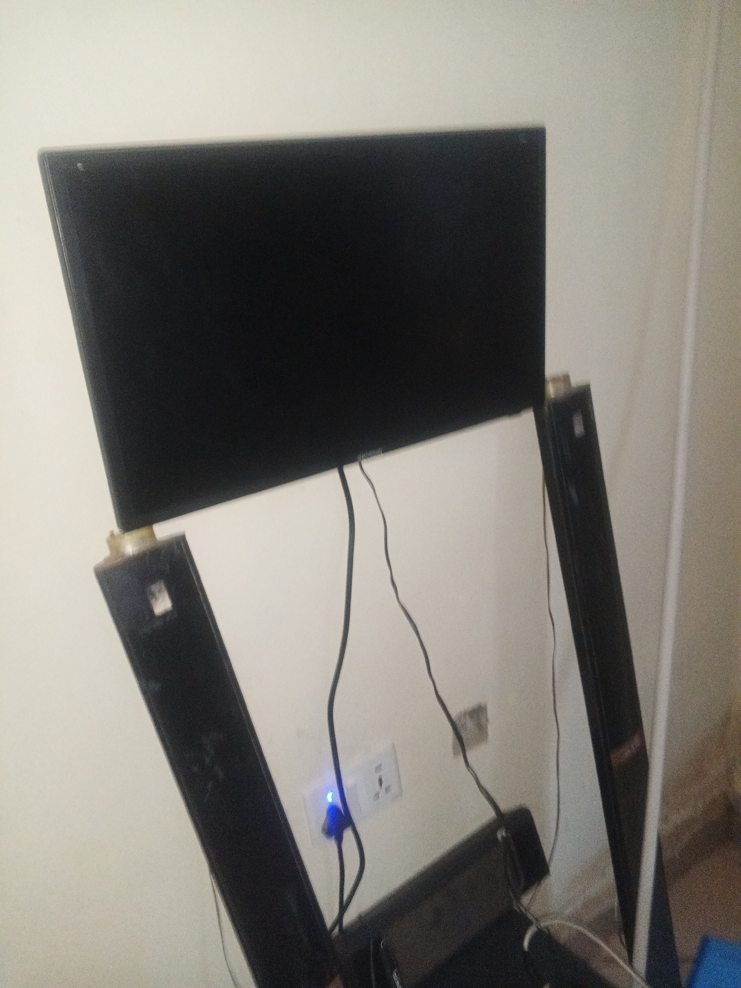 The image shows a flat-screen television set, likely a smart TV given the context of Jalingo, Nigeria, where smart technology is becoming more prevalent. The TV is supported by a black stand, with two vertical components on either side. These stand components are sleek and modern, suggesting a contemporary setup. Below the TV, a black soundbar is visible, indicating an enhanced audio experience. The entire setup is positioned against a plain, light-colored wall, possibly painted plaster.
A power outlet is visible on the wall, with a blue light emanating from a plugged-in device, suggesting the electronics are powered on. Several black cables are neatly organized and connected to the devices, trailing down the wall and behind the soundbar. The lighting in the scene appears to be artificial, and the overall ambiance is domestic and functional. There are no people visible in the image. The scene is static, with no discernible activity taking place beyond the passive display of the television and the powered state of the electronics.
The setting of Jalingo, Nigeria, is a general context for the image, suggesting the location of this electronic setup. The image focuses on the technology and its immediate surroundings, highlighting a modern entertainment system within a typical interior. The details are factual: a TV, stand, soundbar, wall, outlet, and cables. The image does not provide cues about the time of day or weather. No discernible text is present on the objects within the frame.
