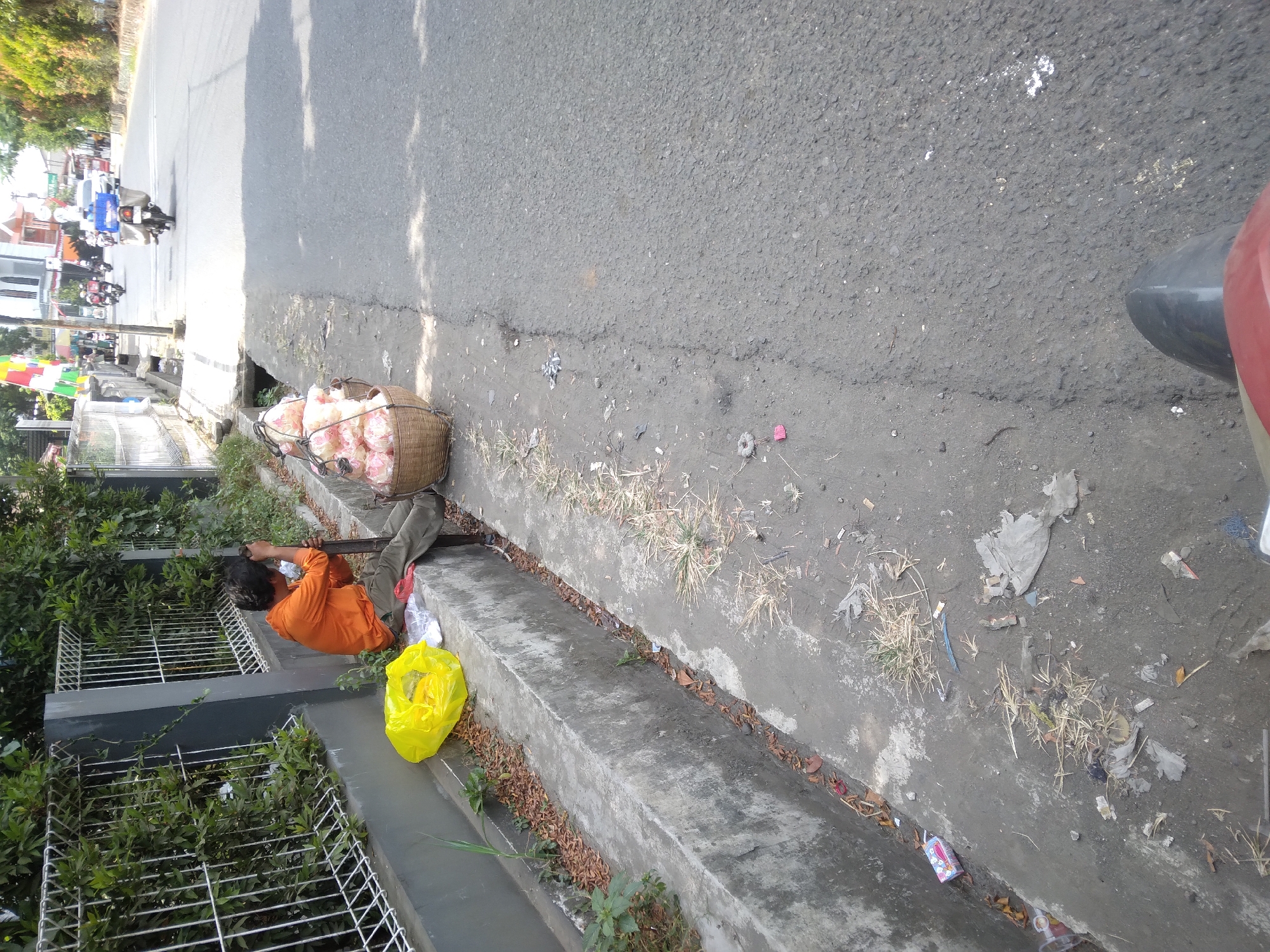 The image captures a street scene in Sokaraja, Indonesia, near a drainage ditch. In the foreground, a person wearing an orange shirt and dark pants is seated on the edge of the concrete drainage ditch, holding a long stick. A yellow plastic bag is placed next to them, along with some white items. To the right of the seated person are two woven baskets filled with light-colored, round objects, likely food items. The drainage ditch is filled with debris, including dry leaves and some green vegetation. Beyond the ditch, the asphalt road is visible, with a motorcycle in the distance carrying a blue container. The environment suggests an urban setting with buildings and trees lining the street in the background. The overall impression is of a vendor resting by the roadside.