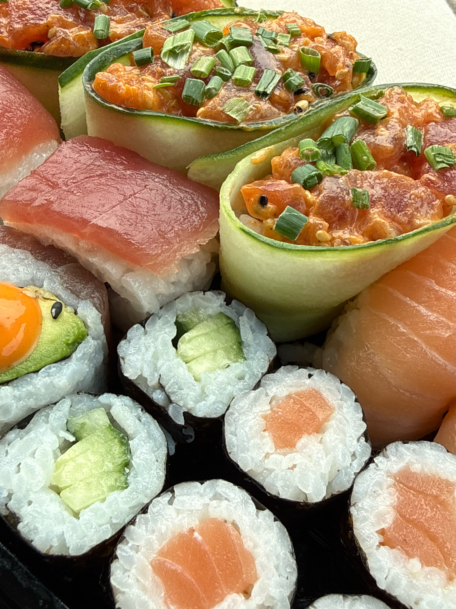 This is a close-up, high-angle, eye-level shot of a platter of assorted sushi. The platter is dark and appears to be made of plastic or a similar material. The sushi pieces are arranged densely and include various types:
* **Poke bowls:** Two distinct servings of chopped raw fish mixed with sauce and topped with chopped green onions. These are contained within rolled slices of cucumber.
* **Nigiri:** Several pieces of nigiri sushi are visible, featuring a slice of red tuna on top of rice, and a piece with a slice of salmon on rice.
* **Maki rolls:** Several cut maki rolls are visible, some with cucumber filling and others with salmon filling. One maki roll appears to have avocado and possibly a piece of orange, perhaps carrot or sweet potato, inside.
The lighting is bright, highlighting the fresh colors and textures of the sushi. There are no people, identifiable landmarks, or indications of a specific location within Herzberg, Germany, in this image. The focus is solely on the food presentation.