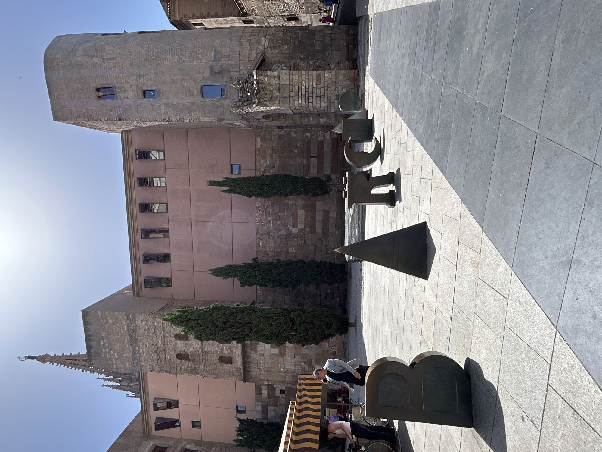 This is a street-level, wide shot of the Plaça de Sant Miquel in Barcelona, Spain, on a sunny day. The foreground is dominated by large, bronze letters spelling out "BARC". To the left, a person wearing a white tank top and dark pants stands next to a bicycle, while a man in a light grey jacket is on his phone. Behind them is a striped awning. Dominating the background are stone buildings, including a tall, cylindrical tower and a larger structure with a pink facade. Several cypress trees are planted in front of the buildings. A few other people are visible in the distance.