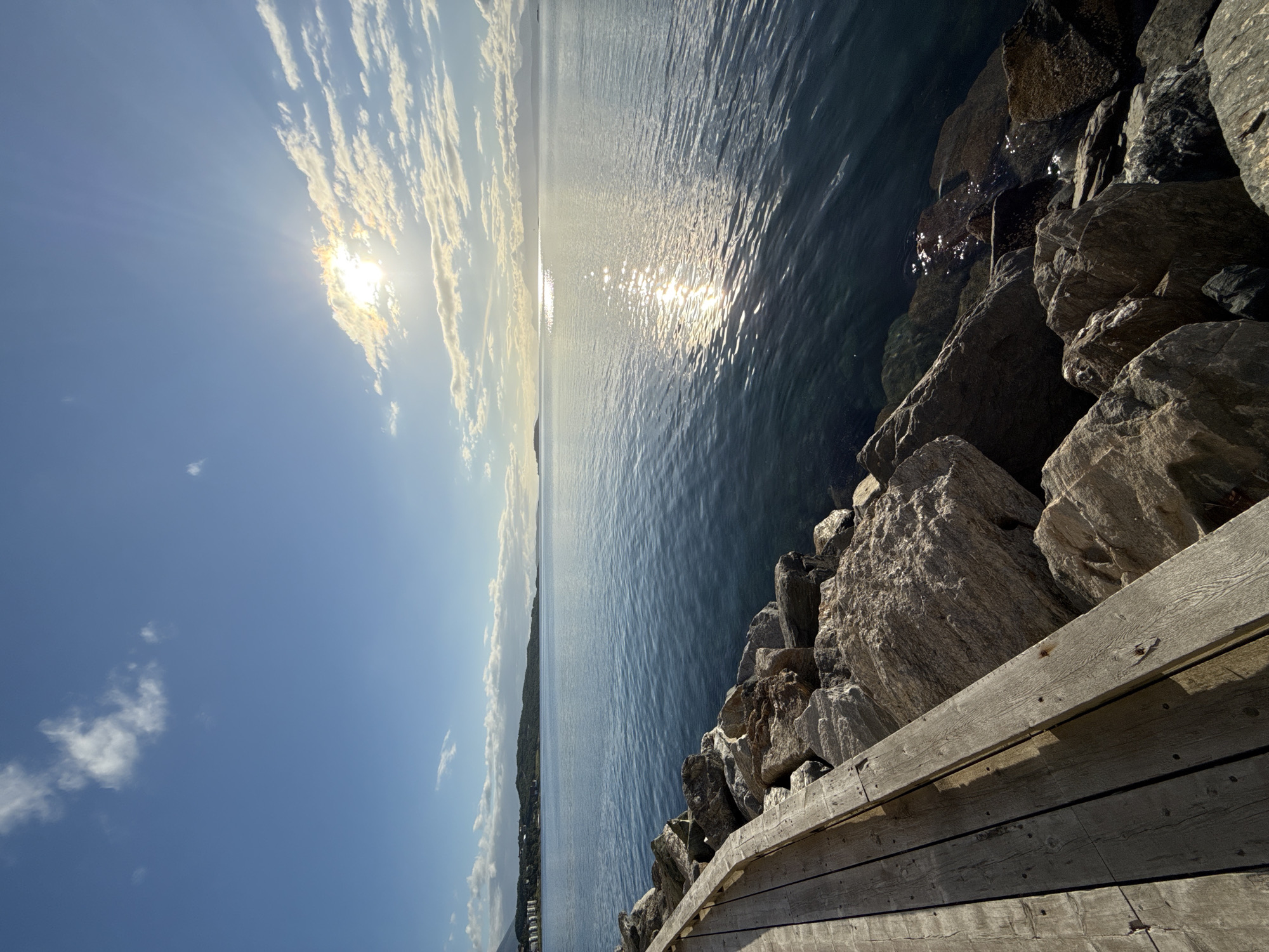 The image depicts a scenic view of the sea with a wooden boardwalk and a rocky shoreline in the foreground. The sun is shining brightly in the sky, casting a reflection on the water. In the distance, a coastline with hills and buildings can be seen. The location is identified as Harstad, Norway. There are no people or animals visible in the image. The scene is peaceful and serene, with the natural beauty of the landscape being the main focus. The boardwalk appears to be made of weathered wood, and the rocks are rough and uneven, suggesting a natural and unmanicured environment. The water is calm and clear, with gentle ripples reflecting the sunlight. The sky is a clear blue with scattered white clouds. The overall impression is one of a tranquil and picturesque coastal setting.