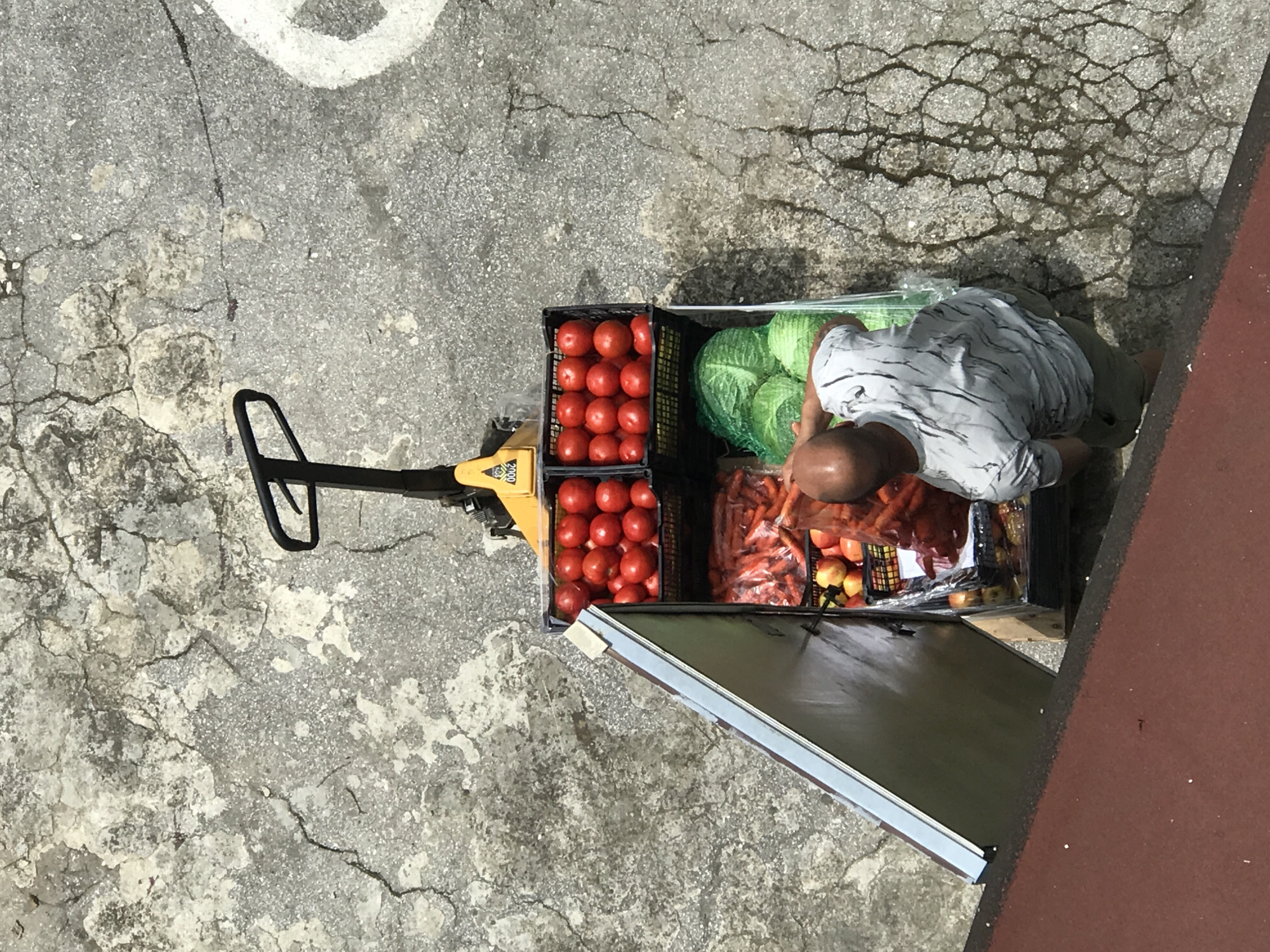 An overhead shot shows a man, from the back and shoulder, bending over a pallet jack loaded with crates of produce. The pallet jack has a yellow handle with the number "0002" visible on it. The crates contain tomatoes, carrots, apples, and cabbages. The man is wearing a light gray t-shirt with a swirled pattern and khaki shorts. He appears to be arranging or packing produce into a plastic bag. The pallet jack and produce are on a cracked, grey concrete surface, possibly a street or sidewalk. A dark red surface is visible in the lower left corner, possibly a building or awning. The scene is set outdoors, under natural lighting. The image context places this scene in Varna, Bulgaria, though the image itself does not contain any visual cues to confirm this specific location.