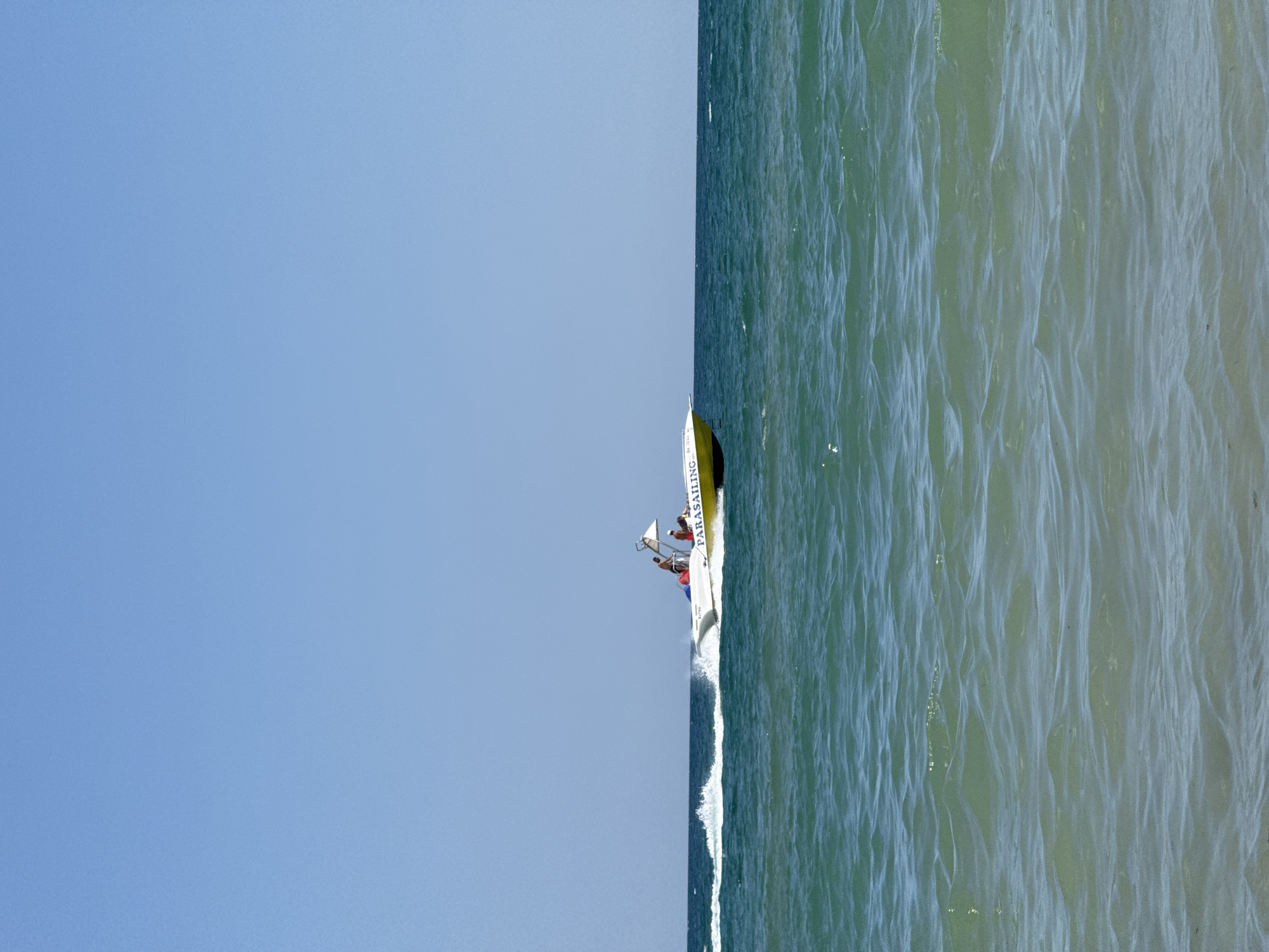 A white and yellow parasailing boat moves left to right across the frame on a calm, teal ocean under a clear blue sky. The boat has two people visible, one wearing a red shirt and the other a white shirt, both standing. The word "PARASAILING" is written in yellow on the side of the boat. The boat is creating a white wake behind it. The image appears to have been taken from a beach in Kranevo, Bulgaria, given the context provided.