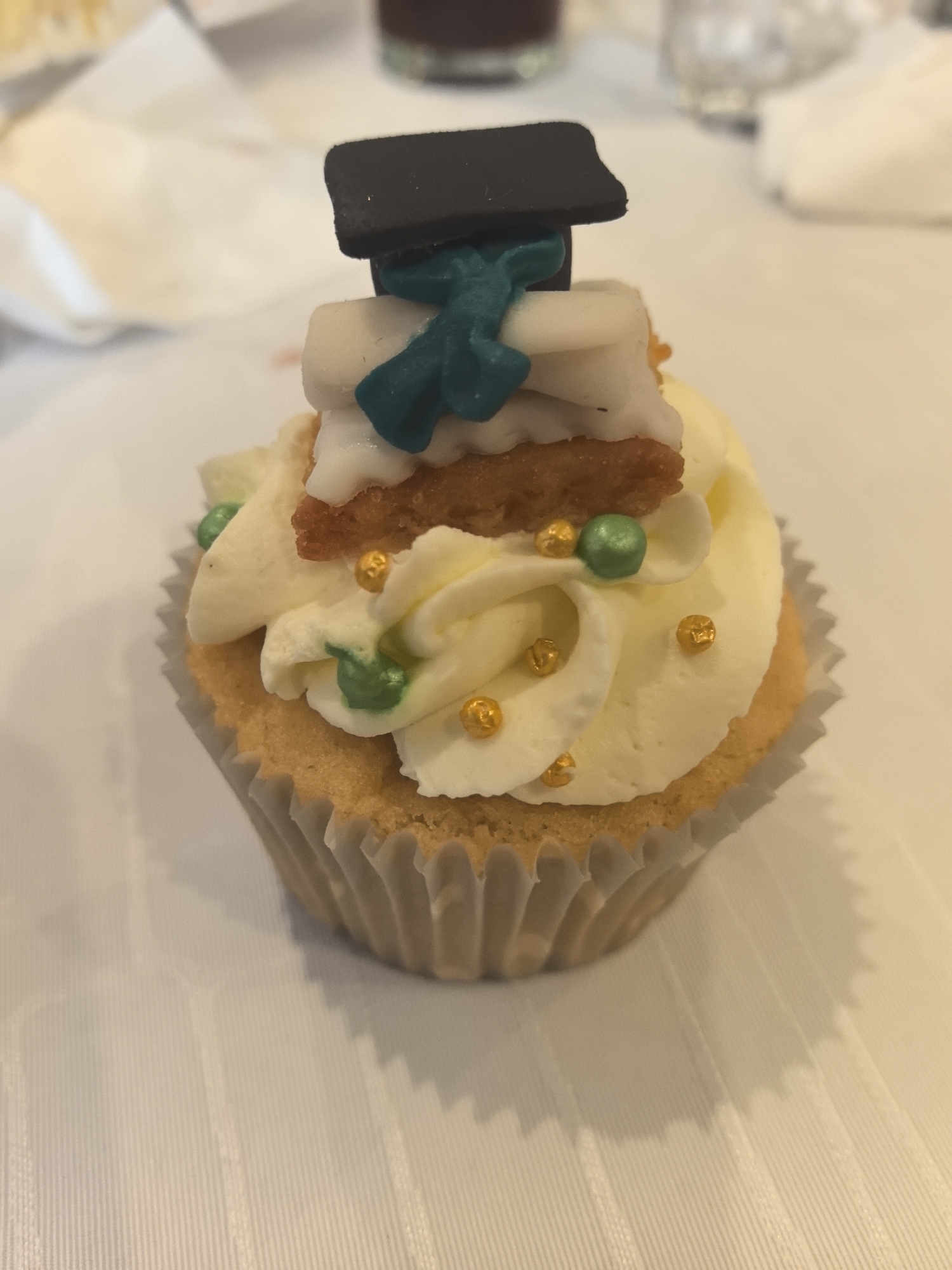 The image shows a cupcake with white frosting in a ridged paper liner. The cupcake is topped with a black square resembling a graduation cap, a teal-colored decoration, and white decorations, as well as small gold and green sprinkles. The cupcake sits on a white, possibly tablecloth-covered surface. There is no information available to determine the specific location in Sofia, Bulgaria, or any other details about the scene.