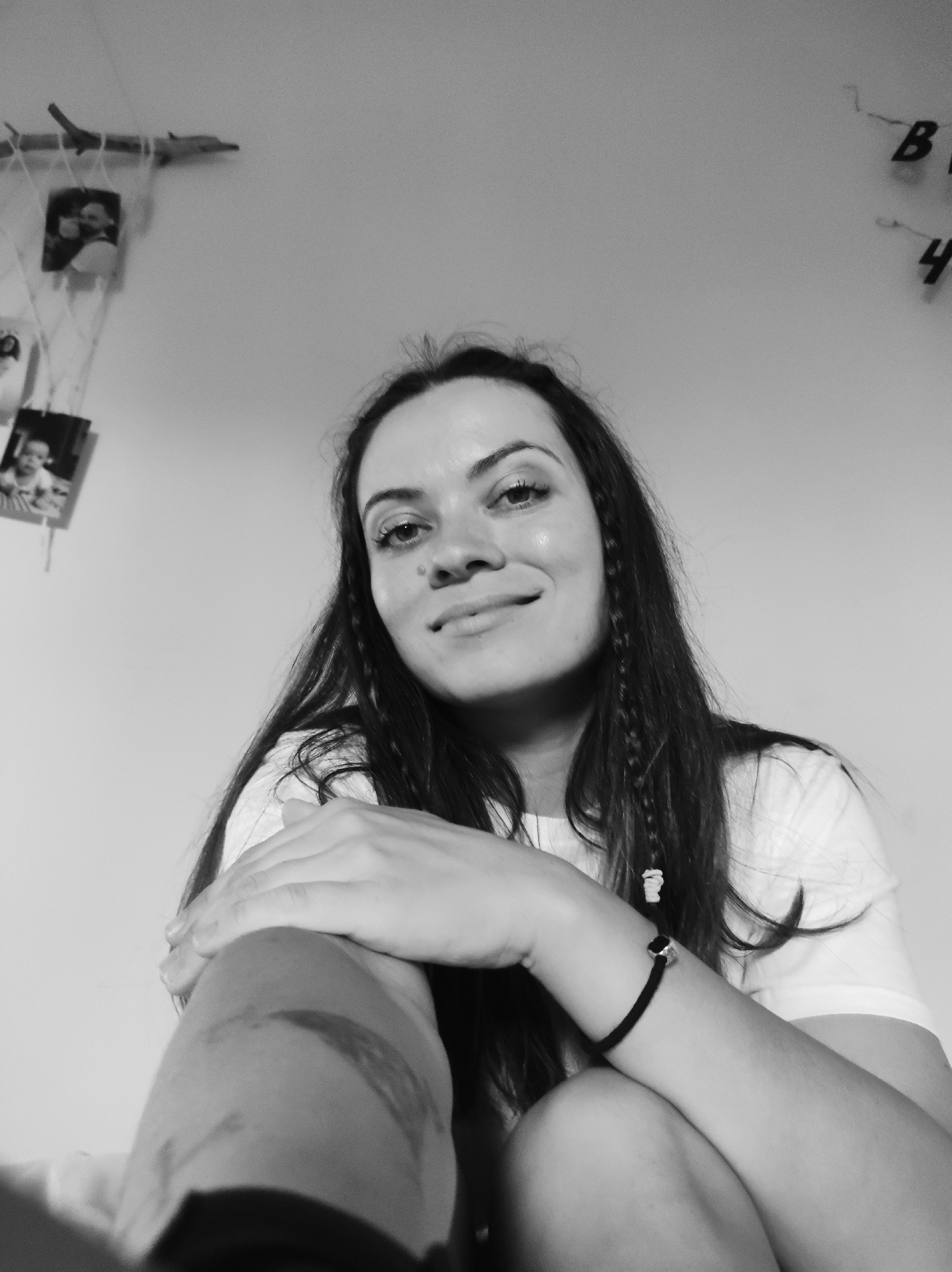 A grayscale, low-angle, medium-close-up photograph depicts a young adult female seated indoors. The woman, with long, dark hair featuring small braids adorned with white beads, looks directly at the camera with a slight smile. Her left arm, wearing a black elastic bracelet with a silver-colored bead, is crossed over her right forearm, which displays several discolored marks. She is wearing a light-colored, short-sleeved t-shirt. The background consists of a plain, light-toned wall. On the upper left side of the wall, a horizontal wooden branch is affixed, from which several small framed photographs are suspended by strings. These photographs appear to show individuals, including a child. On the upper right side of the wall, dark-colored letter-like shapes, specifically 'B' and '4', are visible, attached to the surface. The specific location is Unknown, Unknown.