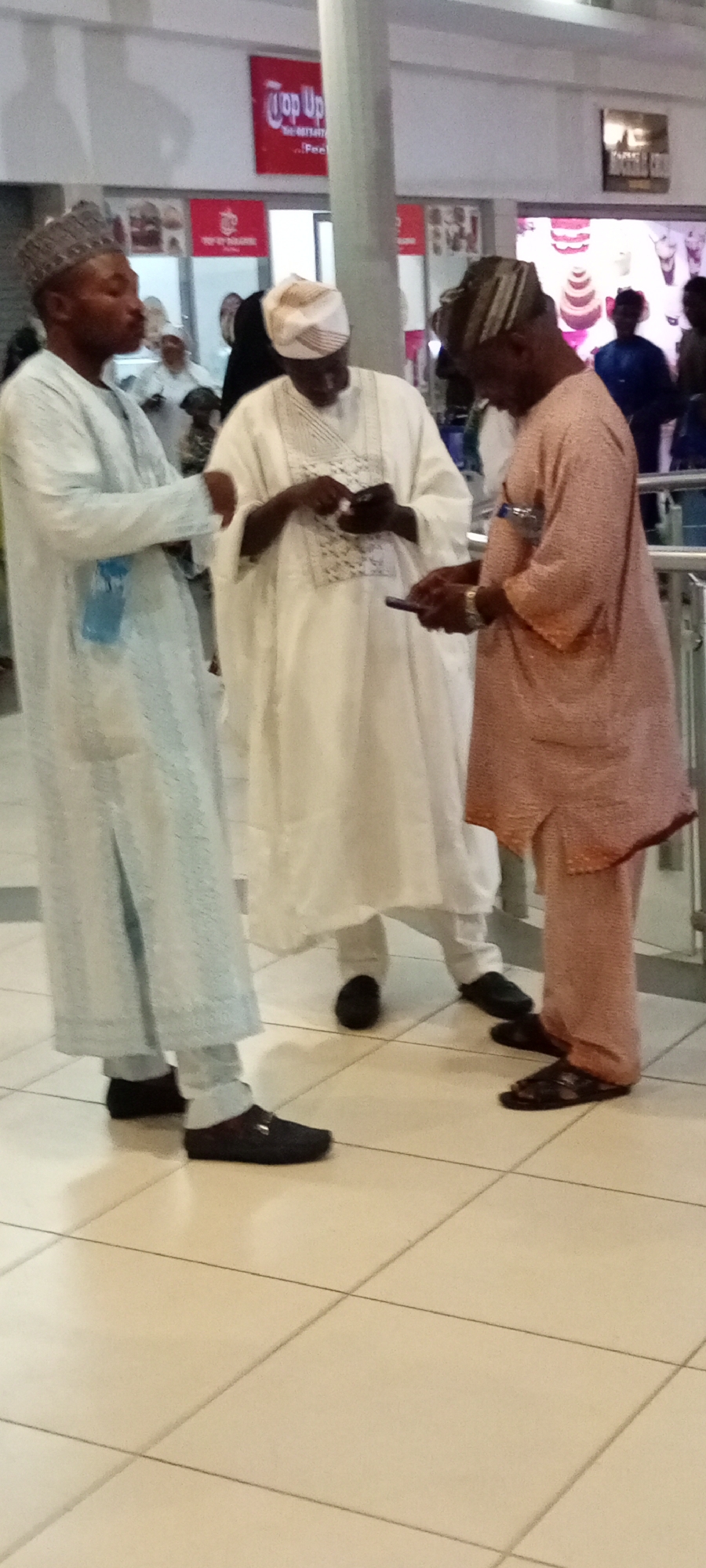 Three adult males are standing on a tiled floor in what appears to be an indoor public space, possibly a shopping mall or transit hub in Gandu Albasa, Nigeria. The man on the left wears a light blue traditional outfit and a matching patterned cap. He is looking down and to his right. The central figure is dressed in a white traditional outfit and cap, and he is holding a mobile phone and looking at it. To the right, another man in a peach-colored traditional outfit and cap is also looking at his phone, which he holds horizontally. His left hand is resting on his thigh. In the background, there are signs, including one that reads "Top Up," and other individuals are partially visible. The lighting suggests it is daytime, and the environment seems clean and well-maintained.