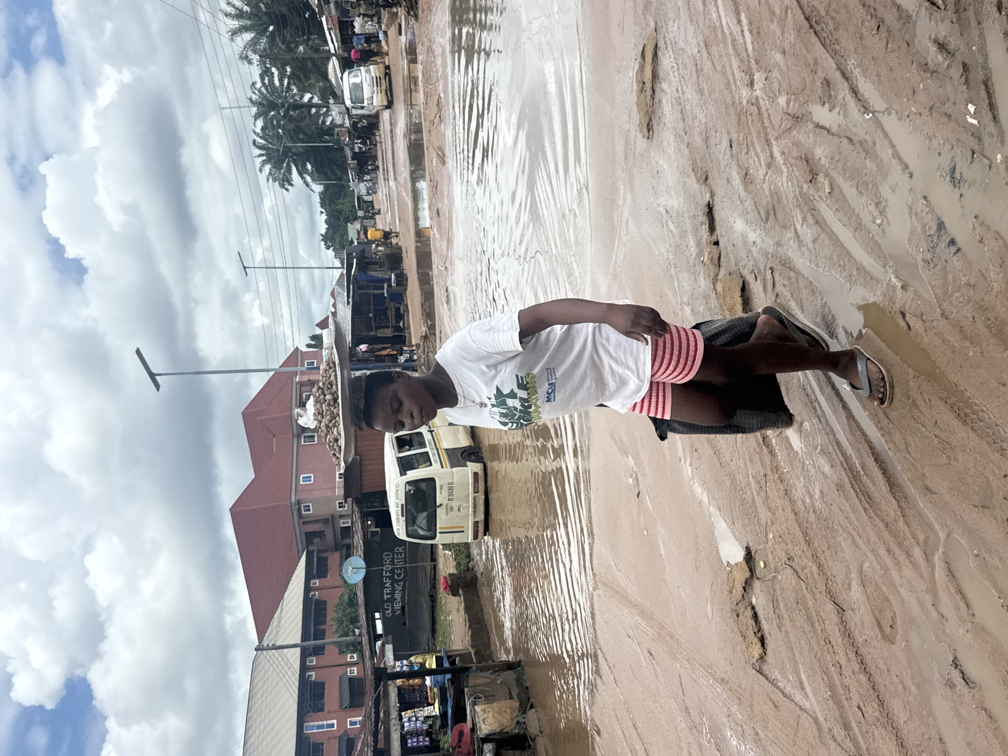 A young woman walks down a muddy, flooded street in Ihiagwa, Nigeria. She carries a tray of goods balanced on her head and a black bag in her left hand. The street is heavily waterlogged, with visible tire tracks and debris. Several buildings line the street, some with red roofs, and a white minibus is visible in the background, partially submerged in water. The sky is cloudy, suggesting recent or ongoing rainfall. The scene depicts the challenging environmental conditions and daily life in the area.