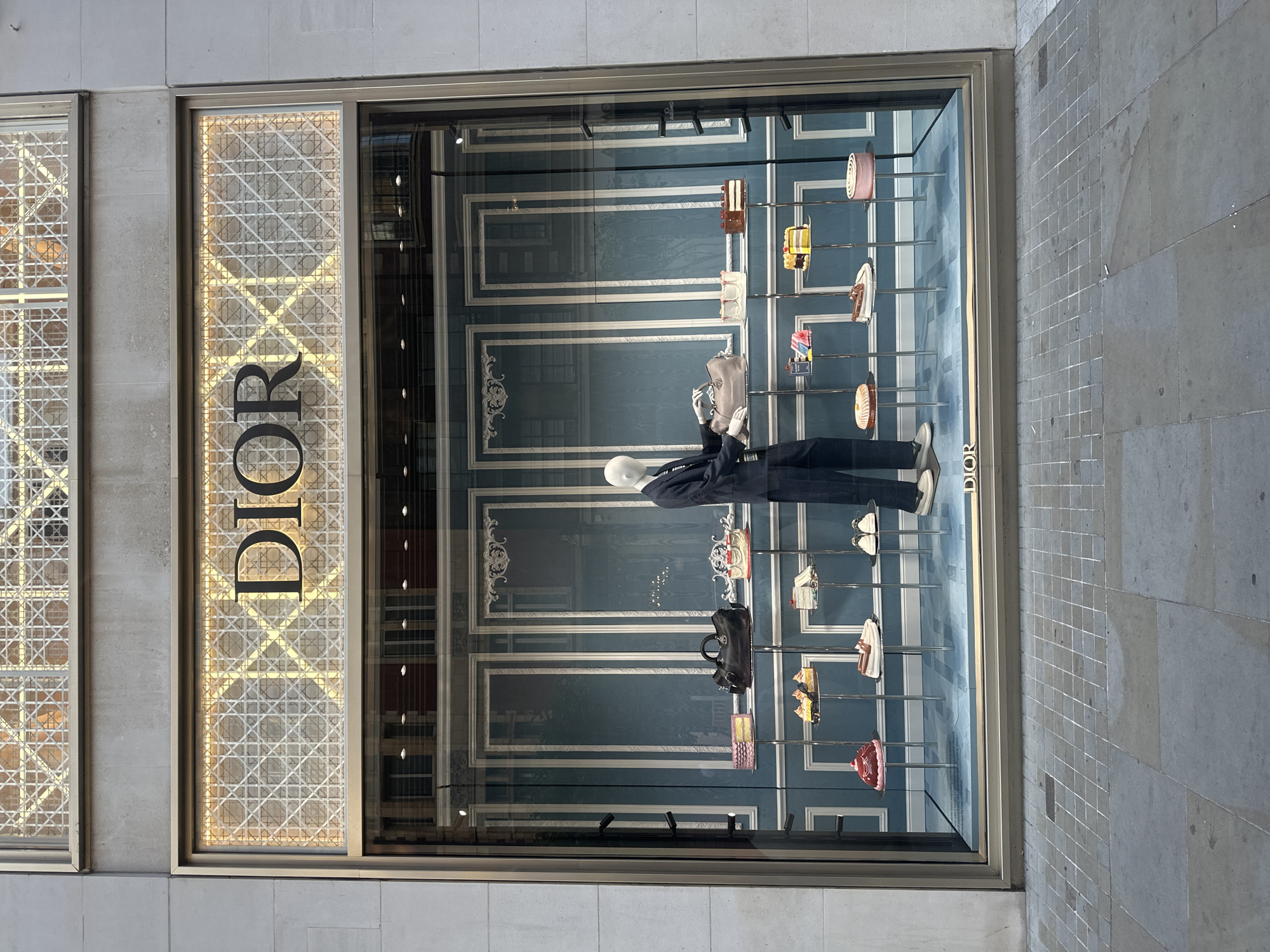 A large, well-lit storefront window displays merchandise from the luxury brand Dior. The window is framed by a textured, pale gray building facade. Above the glass, a decorative metal lattice is illuminated from behind, and the Dior logo is prominently displayed in black lettering against this illuminated background.
Inside the window, the backdrop is a sophisticated teal wall with decorative white molding that resembles paneled walls. Several tiered display stands are arranged, showcasing various items. On the top shelf, a large black handbag is placed next to a pink-tiered cake and a slice of cake. Below this, the shelves hold an assortment of items including: several slices of cake, a handbag in a light beige color, a pair of white loafers, and a pair of white sneakers.
To the right of the display, a mannequin dressed in a dark navy suit with white sneakers is positioned. The mannequin's arms are extended, with one hand holding the light beige handbag. The floor of the display area is a light blue, with a subtle pattern.
The street in front of the storefront is paved with large, rectangular gray tiles, with some white lines indicating seams between them. The overall impression is one of high-end retail display, with a focus on luxury fashion accessories and cake-inspired decorative elements.