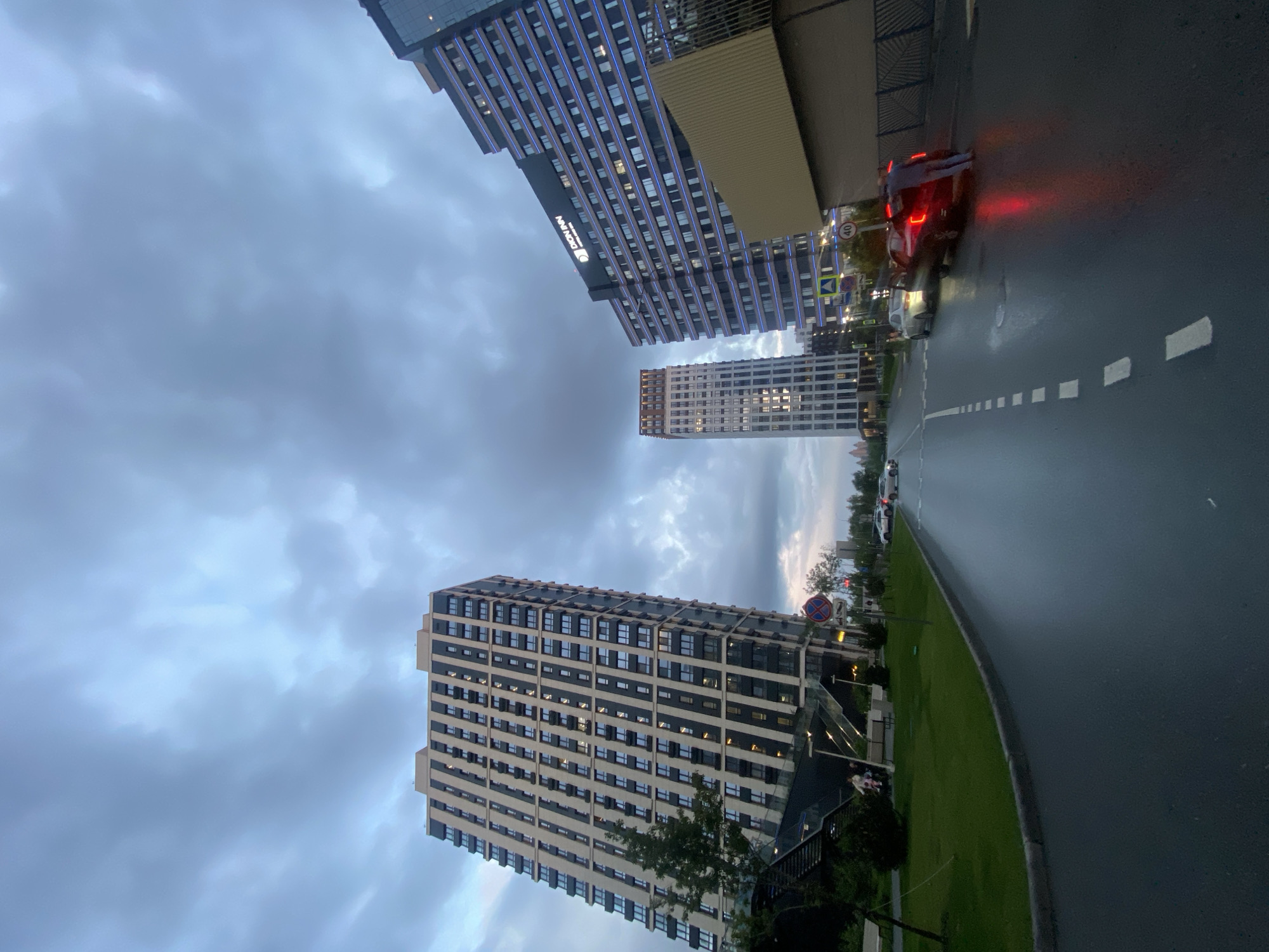 The image, when viewed upright, depicts a modern urban street scene in Kazan, Russia, under an overcast sky. The perspective is low-angle, looking up towards the buildings and the sky, with a wide, wet asphalt road in the foreground and stretching into the middle distance.
On the left side, a prominent, multi-story building features a patterned facade of alternating light and dark horizontal bands, with many windows, some showing interior lights. Further back, another tall building with a lighter facade stands. Towards the upper left, a large, dark-colored modern building with numerous windows, many of which are illuminated with a distinctive blue light, is clearly visible. On its side, the text "DOWINN" is prominently displayed, with "HOTEL & APARTMENTS" visible below it, identifying the building's function.
The road surface is dark and reflective, indicating wetness from recent rain or damp conditions. White dashed lines mark the road. Several vehicles are present: a dark-colored car with bright red tail lights is visible in the middle distance, suggesting it is braking or stopped, with another lighter car behind it. Additional cars are parked further along the road on the right.
A strip of green grass borders the road on the right, interspersed with some trees and bushes. Visible road signs include a circular "40" speed limit sign and a red circular sign with a diagonal line, indicating "No Stopping" or "No Standing."
The sky is filled with varying shades of grey clouds, confirming an overcast day. The combination of diffused daylight and illuminated windows suggests the time of day is late afternoon or early evening, likely dusk. The scene captures a contemporary city environment during a cloudy, possibly rainy, period.