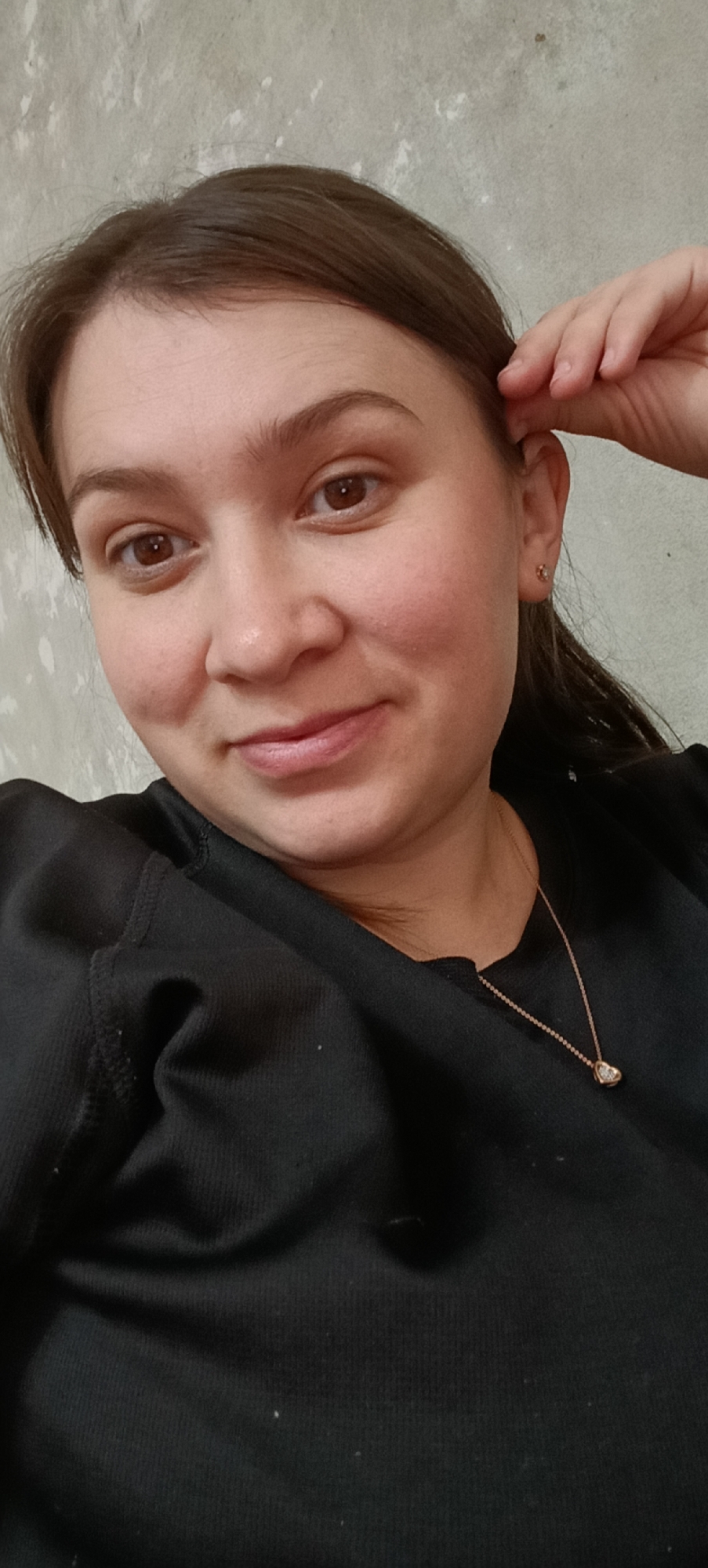 This is a selfie of a young woman with brown hair and brown eyes. She is wearing a black shirt and a delicate gold necklace with a small diamond pendant. She is resting her right hand on her head. The background is a textured, light gray wall. The image was taken indoors, likely in a home or an apartment, given the wall texture and personal setting. The location information provided "Хохлы, Russia" suggests this is where the photo was taken.