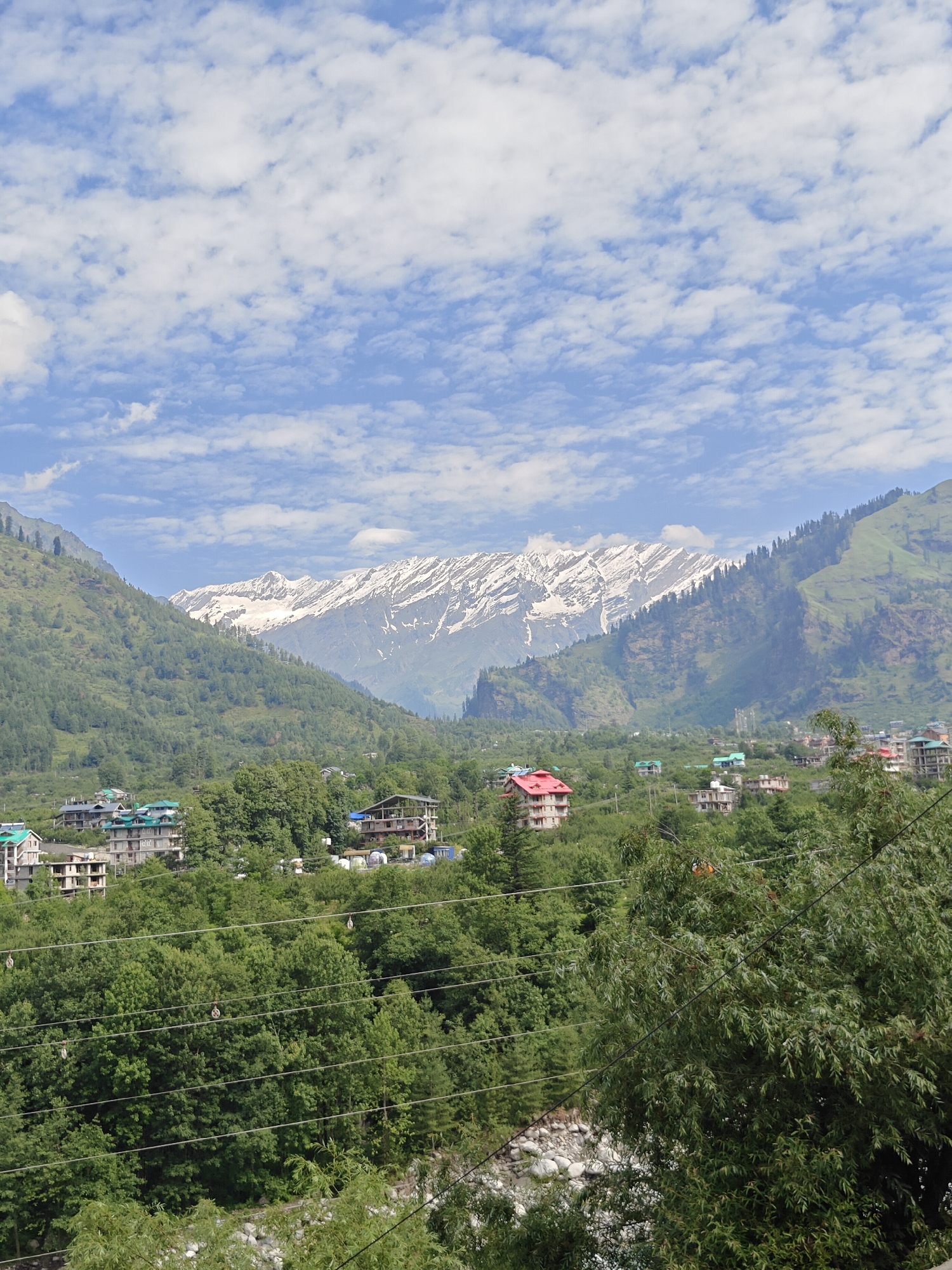 The image depicts a scenic mountainous landscape with a village nestled in the valley. The foreground is dominated by lush green trees and a section of a riverbed with large stones. Several power lines stretch horizontally across the mid-ground. Beyond the trees, a collection of buildings, possibly hotels or houses, are visible, characterized by varied roof colors, including green and red, and architectural styles. In the background, imposing mountains rise, with their peaks covered in snow. The sky above is a bright blue, dotted with scattered white clouds. The overall impression is one of a serene, natural environment interspersed with human habitation, likely situated in or near Kulang, India, given the presence of snow-capped mountains and verdant valleys, consistent with the region. There are no people or animals clearly visible in the frame.