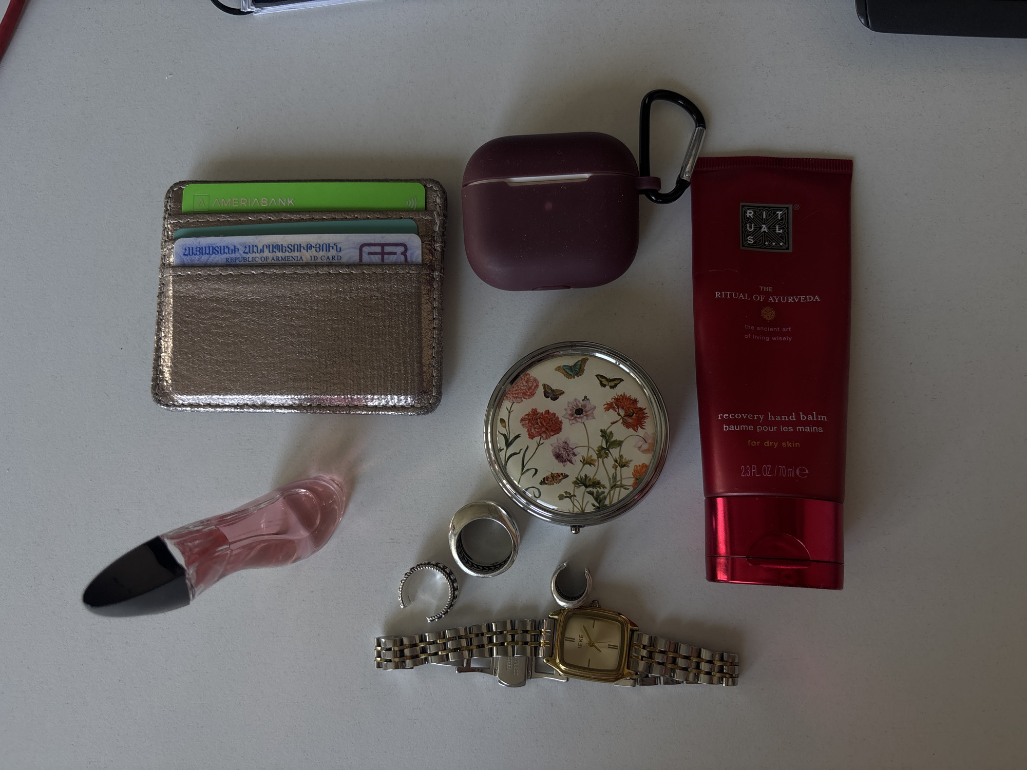 A flat lay photograph features various personal items arranged on a light-colored surface. At the top left, a metallic wallet with cards is visible, including one from "AMERIABANK" and an Armenian ID card. Next to the wallet, there's a burgundy-colored AirPods case with a carabiner clip. Further right, a tube of red "RITUAL OF AYURVEDA" recovery hand balm is positioned.
Below the AirPods case is a round compact mirror with a floral design. A small perfume bottle, pink with a black cap, lies near the wallet. The rings are scattered near the bottom center. A silver and gold watch is laid out horizontally.
The location cannot be determined from the image itself. Additional context or information would be required to identify the specific location in Yerevan, Armenia.