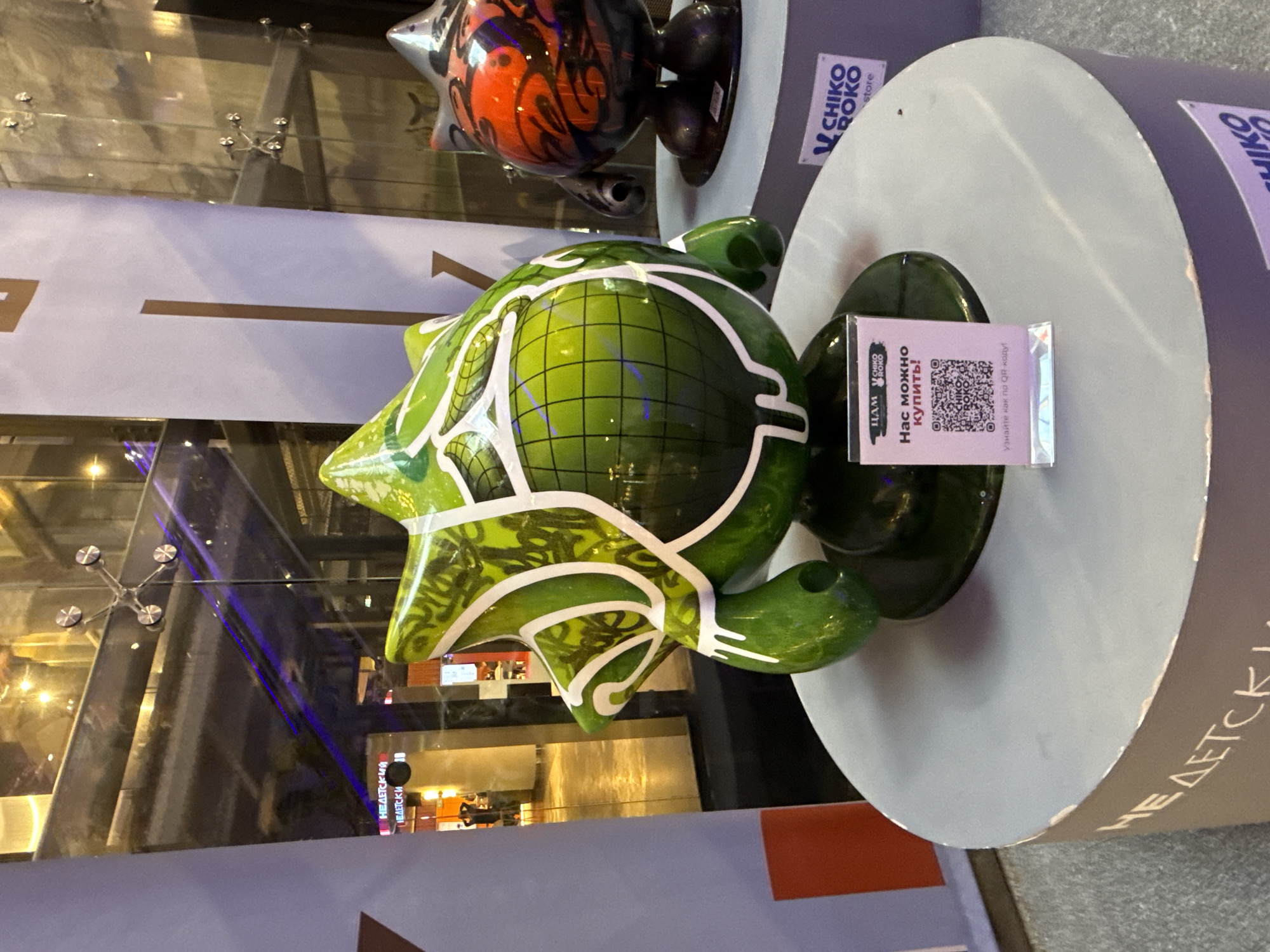 This image captures two stylized art sculptures displayed within a modern indoor commercial or exhibition space, likely a shopping mall, in Moscow, Russia.
The primary subject is a large, glossy green sculpture, approximately waist-height, featuring a complex, multi-pointed, star-like upper section and a spherical body adorned with a dark grid pattern. White, abstract outlines and patterns decorate its green surface. This sculpture rests on a cylindrical, matte grey pedestal. A small, translucent pink sign with white text is placed on the pedestal, displaying the logos "ЦАМ" and "CHIKO ROKO" at the top. The Russian text reads "НАС МОЖНО КУПИТЬ!" (You can buy us!) followed by "Узнайте как по QR-коду!" (Find out how via QR code!), above a printed QR code.
To the right of the green sculpture, a second, smaller sculpture, predominantly red and black with abstract patterns, is partially visible, also situated on an identical grey cylindrical pedestal. A label affixed to its pedestal displays the text "V CHIKO ROKO store".
The background consists of large, clear glass panels, structurally held by spider-fitting hardware, reflecting interior artificial lights, architectural details, and several indistinct human figures. A bright blue linear light reflection traverses the glass horizontally. Beyond the glass, a well-lit interior space with warm lighting and polished floors is visible, indicating a public setting with pedestrian traffic. To the far left, a tall, white vertical banner features partially visible Russian text, including "НЕДЕТСКИЙ" (Adult/Not for children), rendered in white against a pale background with a brown vertical stripe. The overall scene suggests a contemporary art display within a bustling public venue.