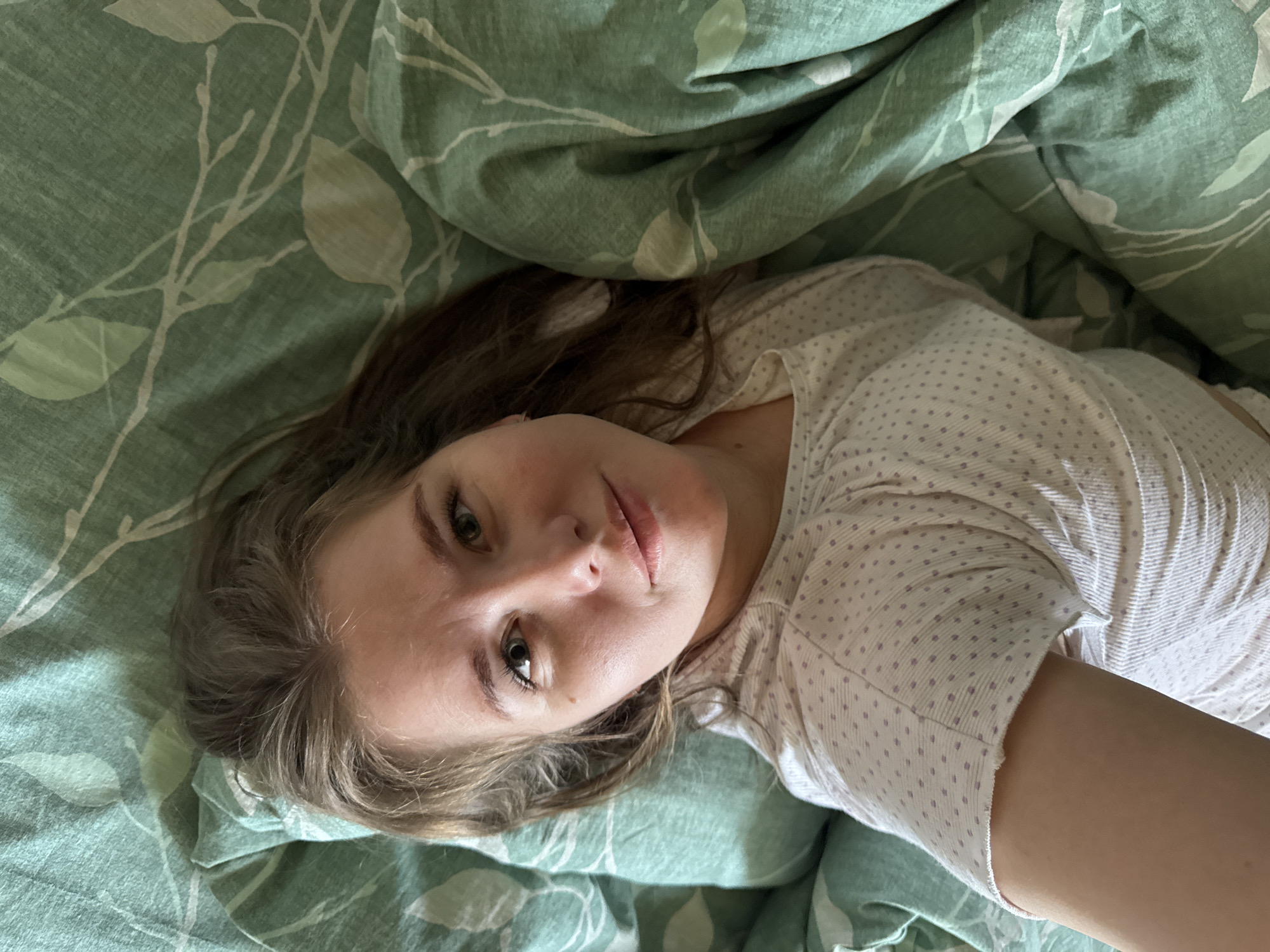A woman lies on a bed in what appears to be a private residence. She has fair skin, light brown hair, and green eyes. She is wearing a white short-sleeved shirt with small purple dots. The bed is covered with green bedding featuring a leaf pattern. The location is Ферма, Russia, inferred by the presence of the location in the user prompt.