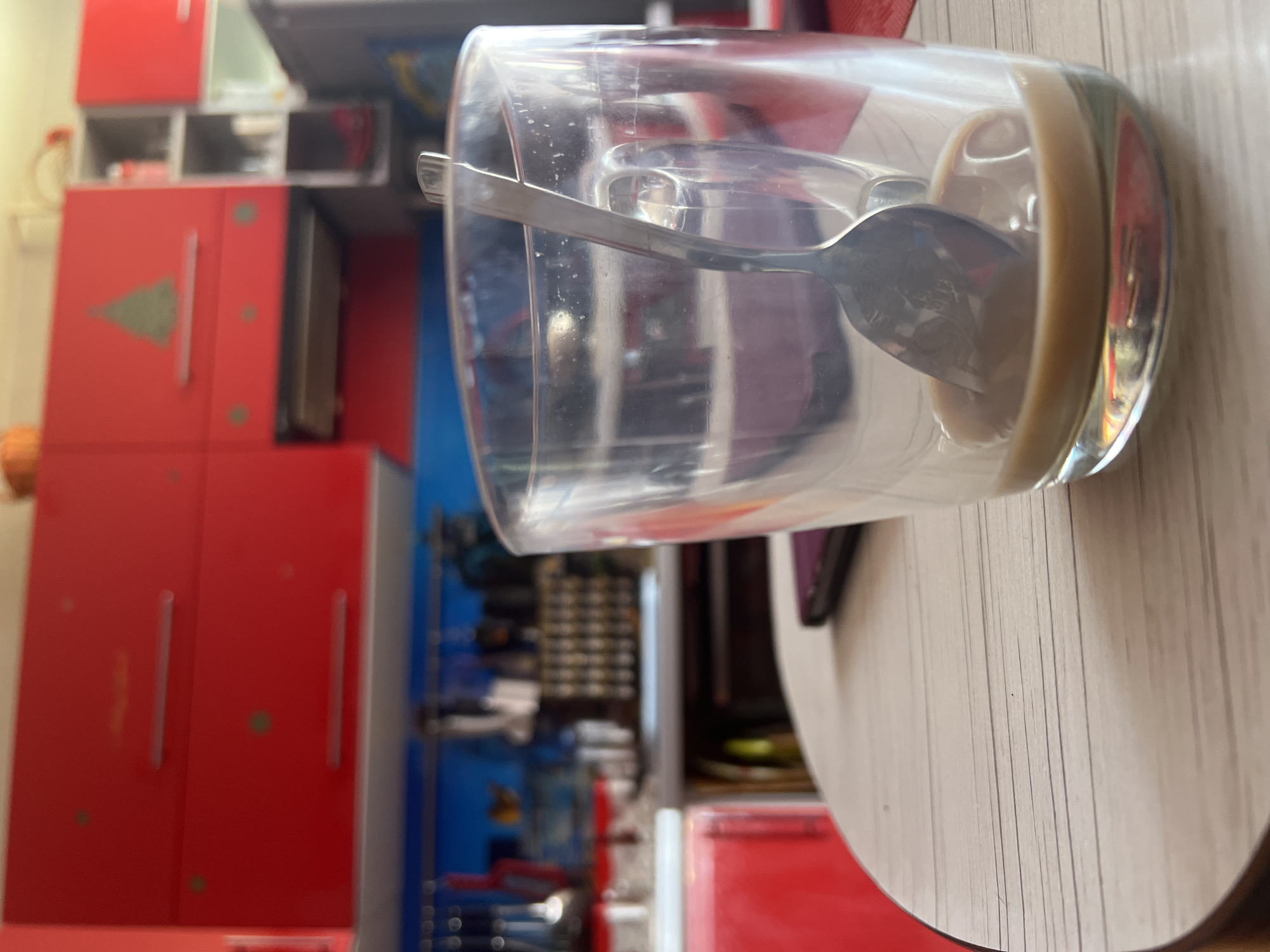 A close-up, tilted, indoor shot shows a clear glass containing a spoon and a small amount of tan liquid, possibly coffee. The glass sits on a light-colored, round table with a wood-grain pattern. The background is a cluttered kitchen, with red cabinets, some decorated with a green tree emblem. A blue section is visible behind some shelving. Tools and miscellaneous objects are also seen in the background. The location, Noviy, Russia, is not definitively discernible from the image's content. No people are visible.
