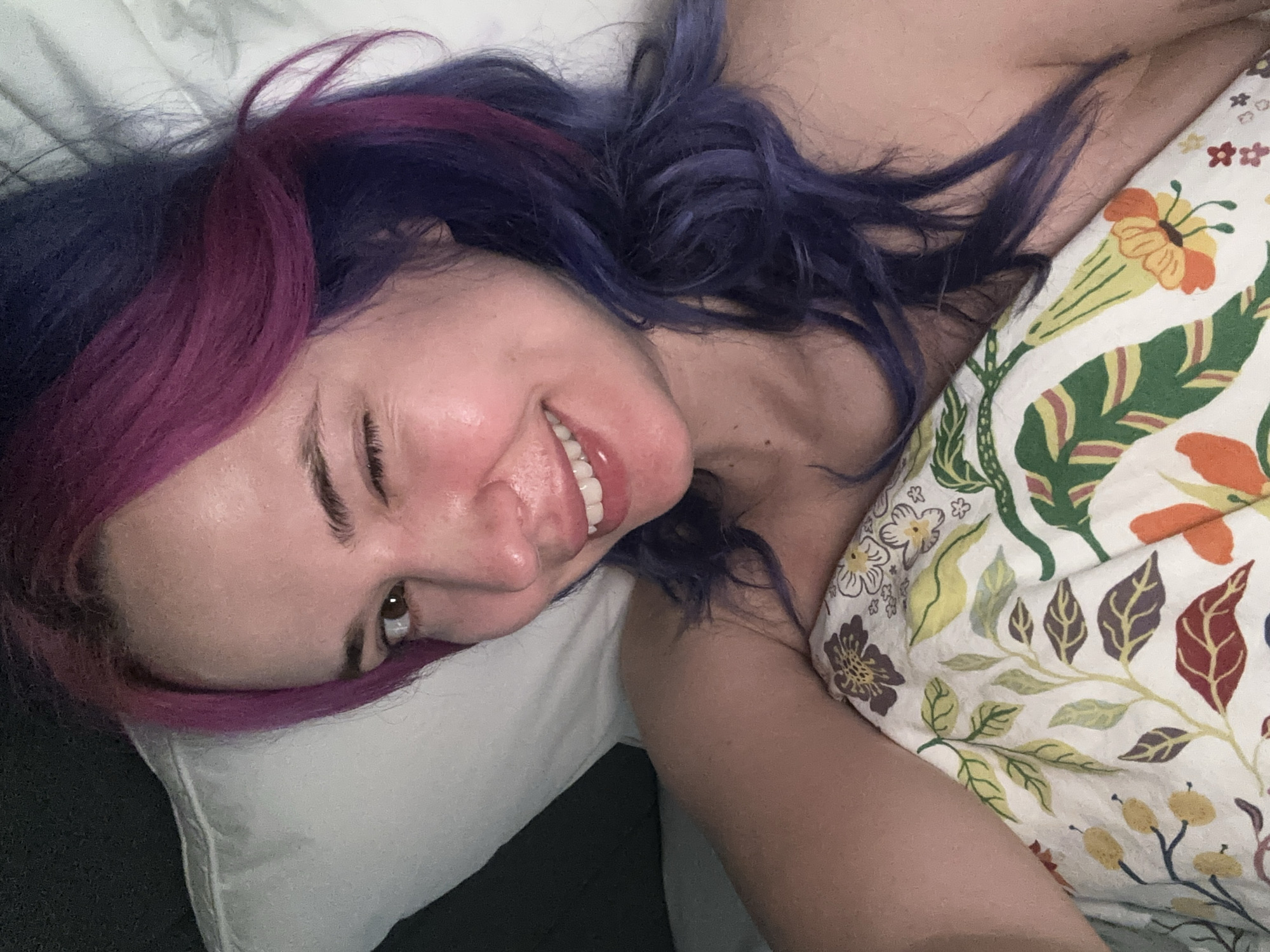 A fair-skinned person with purple and blue hair is lying on a bed, smiling. They are positioned on a white pillow and partly covered by a duvet with a floral pattern. The photo gives no indication that they are located in the city of Zagreb, Croatia.