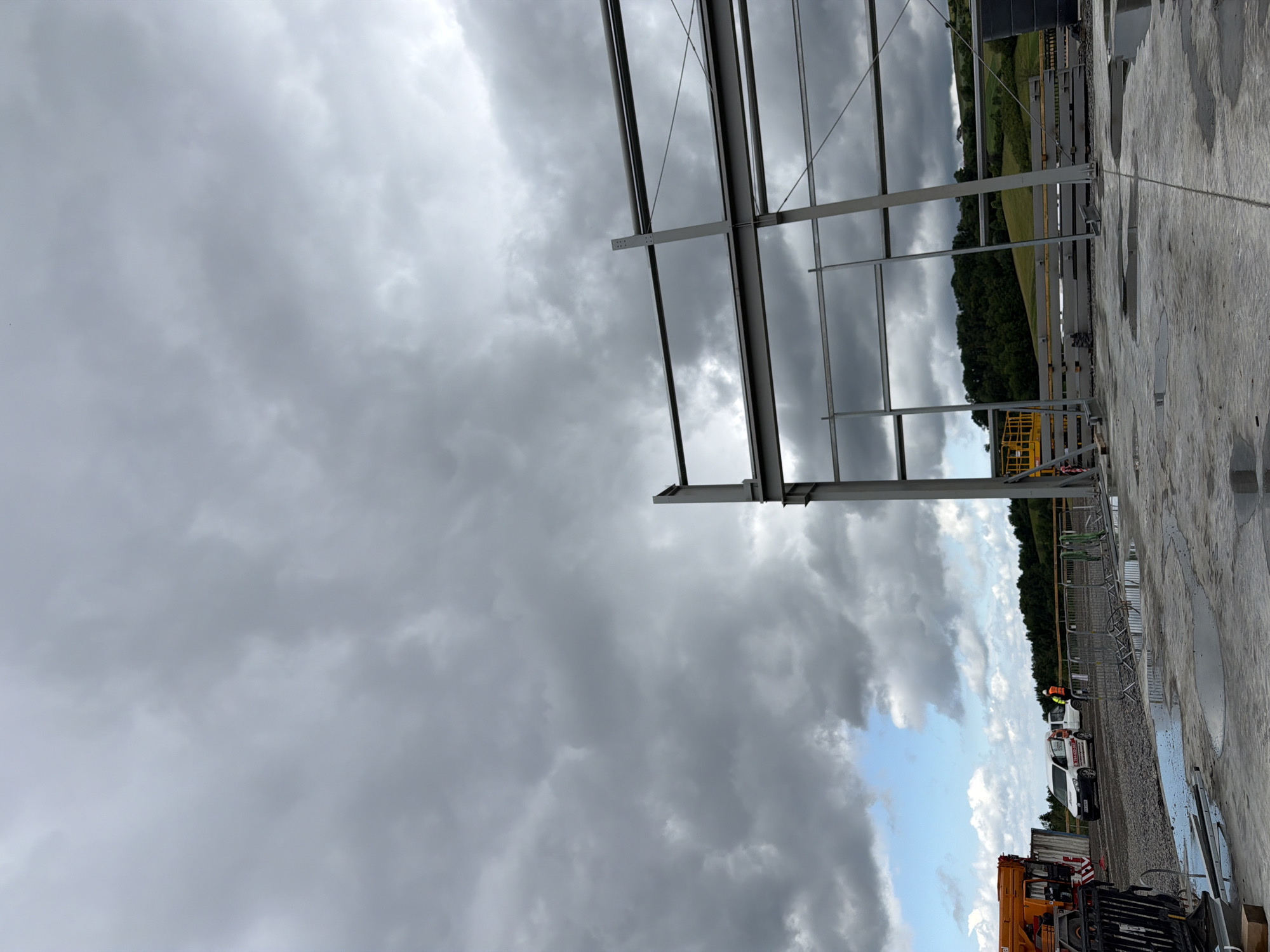 The image depicts a construction site under a cloudy sky. In the foreground, a concrete surface is wet with puddles. To the right, a portion of a large steel structure, likely part of a building frame, is visible. In the mid-ground, a crane truck with an orange boom is parked, next to a white van with signage and a white utility vehicle. A person wearing a high-visibility vest stands near the utility vehicle. Temporary fencing is set up across the area. The background consists of a treeline and rolling hills under a dramatic, overcast sky. The scene suggests ongoing construction activity in an outdoor, rural or semi-rural environment. While the image itself does not provide specific geographical identifiers, if the location is Stróża, Poland, it indicates construction in that area.