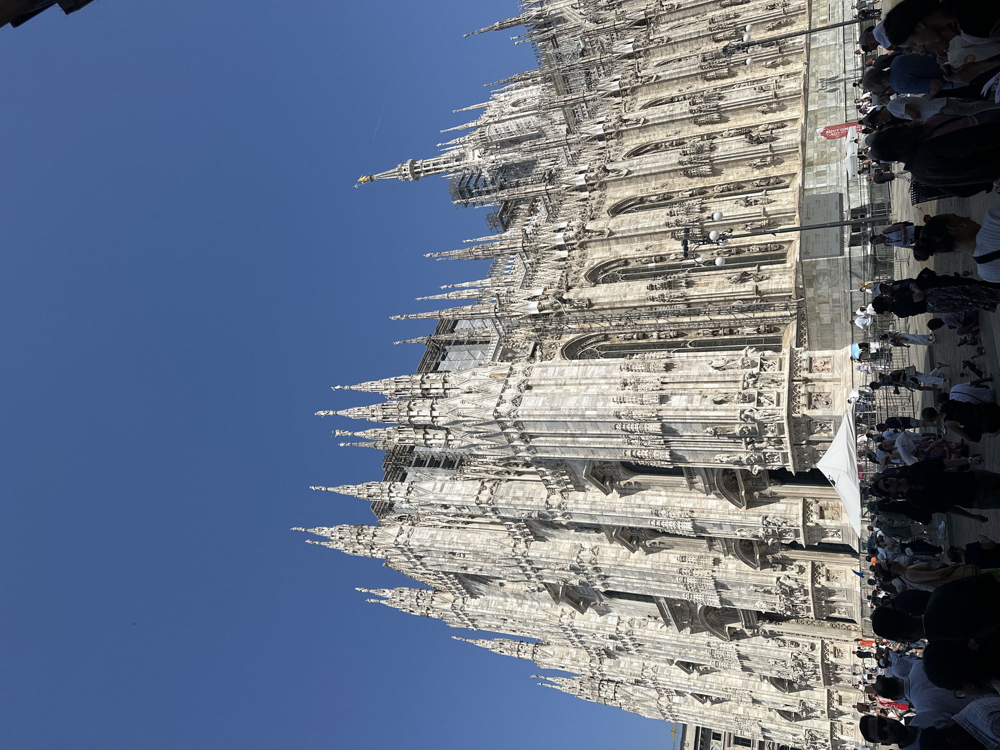 An exterior, low-angle, full shot captures a clear blue sky over the intricate facade of the Milan Cathedral, also known as the Duomo di Milano, located in Milan, Italy. The Gothic cathedral, constructed of white marble, displays numerous spires, statues, and ornate carvings. Some sections of the cathedral show scaffolding, indicating ongoing restoration work. A crowd of tourists and locals are gathered in front of the building, some walking, others standing, and a few are interacting. A white tent is set up near the entrance. Streetlights and a trash can are also visible. The overall scene depicts a sunny day at a prominent historical landmark.