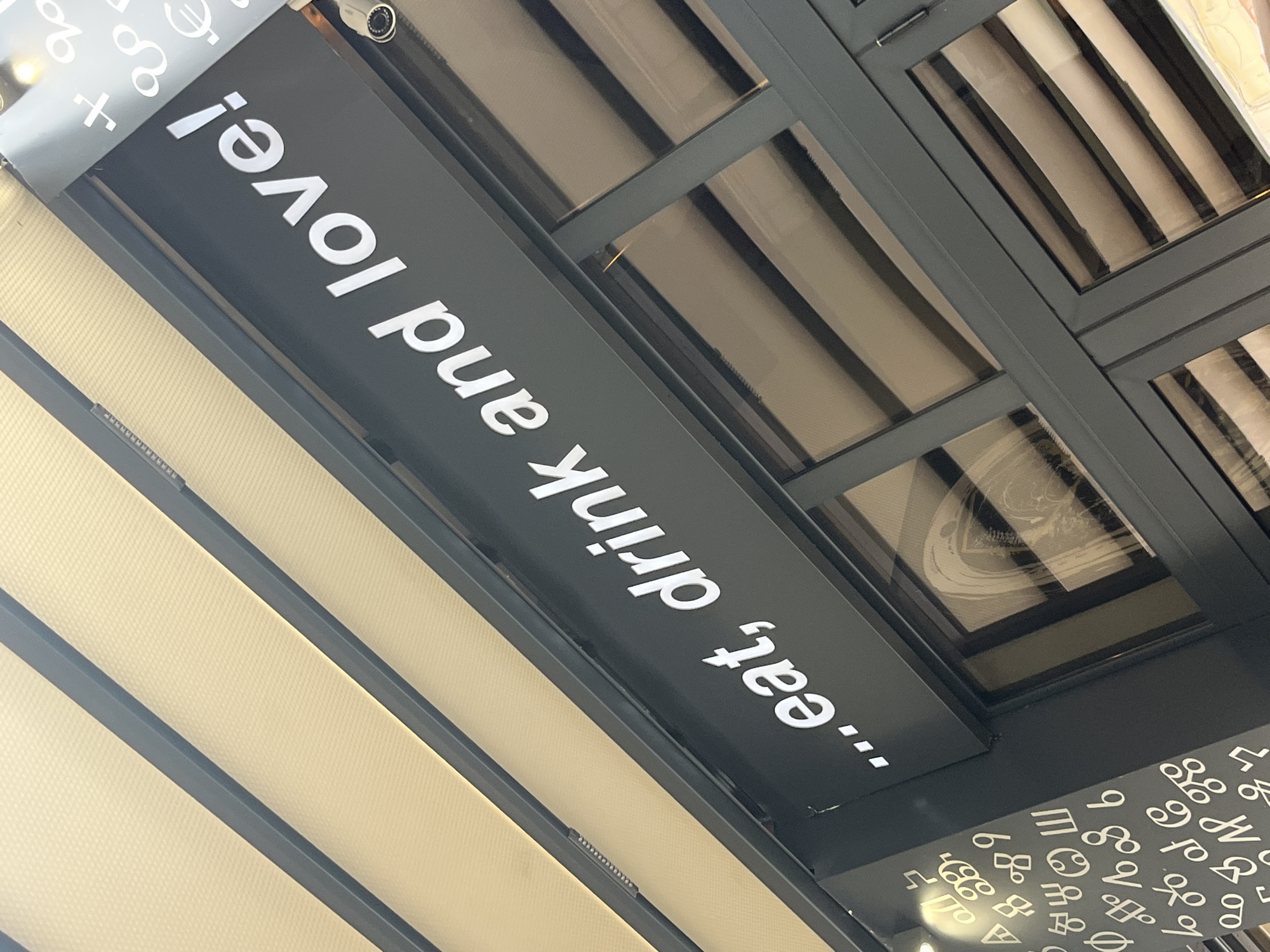 A low-angle shot shows a black sign with white text reading "...eat, drink and love!" The sign is mounted beneath a retractable awning with light-colored slats. Above the awning, the corner of a grid-patterned skylight or window is visible. The sign also features astrological symbols. The location is Plovdiv, Bulgaria.