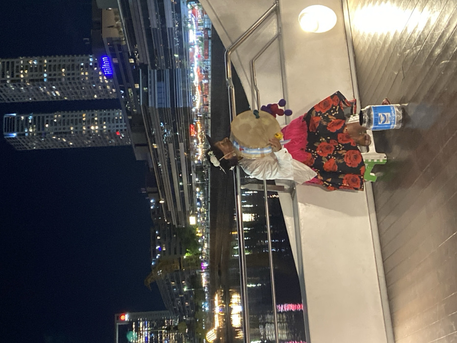 The image depicts a nocturnal scene in Puerto Madero, Buenos Aires, Argentina. In the foreground, on a dark wooden deck, a person wearing traditional attire is seated on a small, light-colored stool, facing right. This individual, likely a woman, is dressed in a white long-sleeved blouse, a fuchsia or pink layered skirt, and a dark skirt featuring a prominent red floral pattern (roses). She wears a dark hat adorned with white flowers and holds a round, hand-held drum with a light-colored head, which is decorated with blue and white ribbons and colorful pom-poms. Directly in front of her on the deck is a clear plastic jar or bottle with a blue label, partially filled with what appears to be coins, functioning as a collection receptacle.
A white railing with a metallic top bar separates the deck from a dark body of water, likely a canal, which reflects numerous city lights. In the background, the illuminated skyline of Puerto Madero rises. On the right, two prominent modern high-rise buildings with many lit windows are visible, and the "Hilton" hotel logo is illuminated in blue letters on a building below them. Further left across the water, another tall, modern building with a red light at its peak and a green light lower down is visible, alongside an industrial crane (likely a historical crane of Puerto Madero). The far bank of the canal features a series of illuminated buildings and establishments, with specific signs like "BigMamma" partially discernible. A light-colored boat or structure with internal lighting is present on the water to the far left.