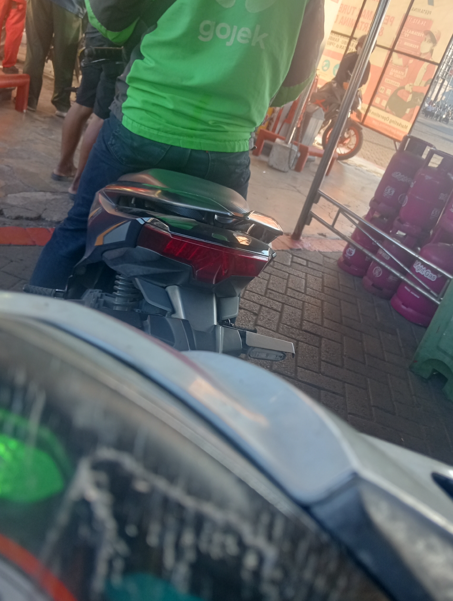 The image shows a close-up view of the rear of a dark-colored motorcycle. The rider, wearing a green jacket with "gojek" written on it and blue jeans, is seated on the motorcycle. The motorcycle has a red taillight. In the background, several pink propane gas cylinders are visible. The ground is paved with interlocking bricks. The scene appears to be outdoors, possibly in an urban setting in Sawotratap, Indonesia, given the presence of the Gojek driver and the gas cylinders, which are commonly seen in such locations.