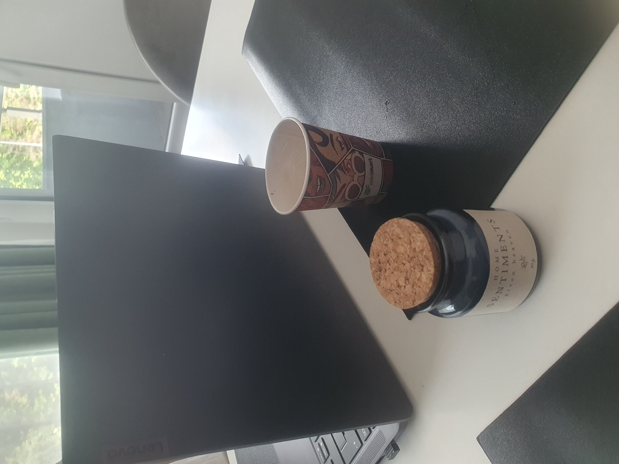 The image shows a table with a laptop (brand: Lenovo), a paper cup, and a small jar labeled "HOME SENTIMENTS Green heaven". The table has a white and black placemat. The location cannot be determined to be in Plovdiv, Bulgaria from the provided image. There are no people visible in the image.