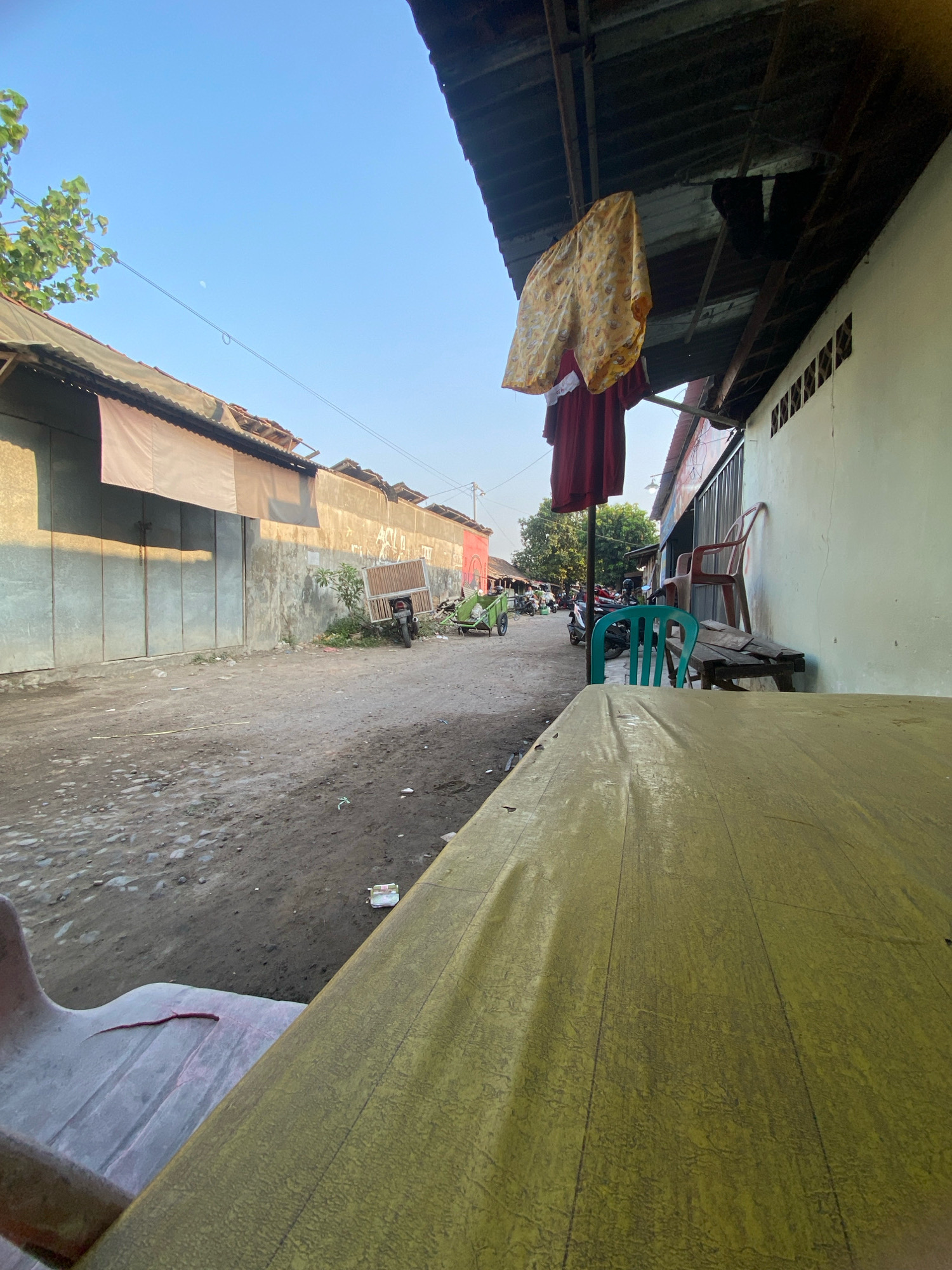 The image depicts a street scene in Gurah, Indonesia. In the foreground, a yellow-covered table is positioned, with a portion of a pink plastic chair visible to the left. The street itself is unpaved and dusty, with scattered debris. On the left side of the street, a large building with metal sliding doors is situated. Further down the street, several motorcycles are parked, and a green cart is loaded with items. On the right side of the street, a light-colored building with a corrugated metal roof runs along the side. Laundry, including a pair of yellow shorts and a red garment, is hanging on a line under the roof. Two plastic chairs, one green and one red, and a wooden bench are placed near the building. The sky is clear and blue.