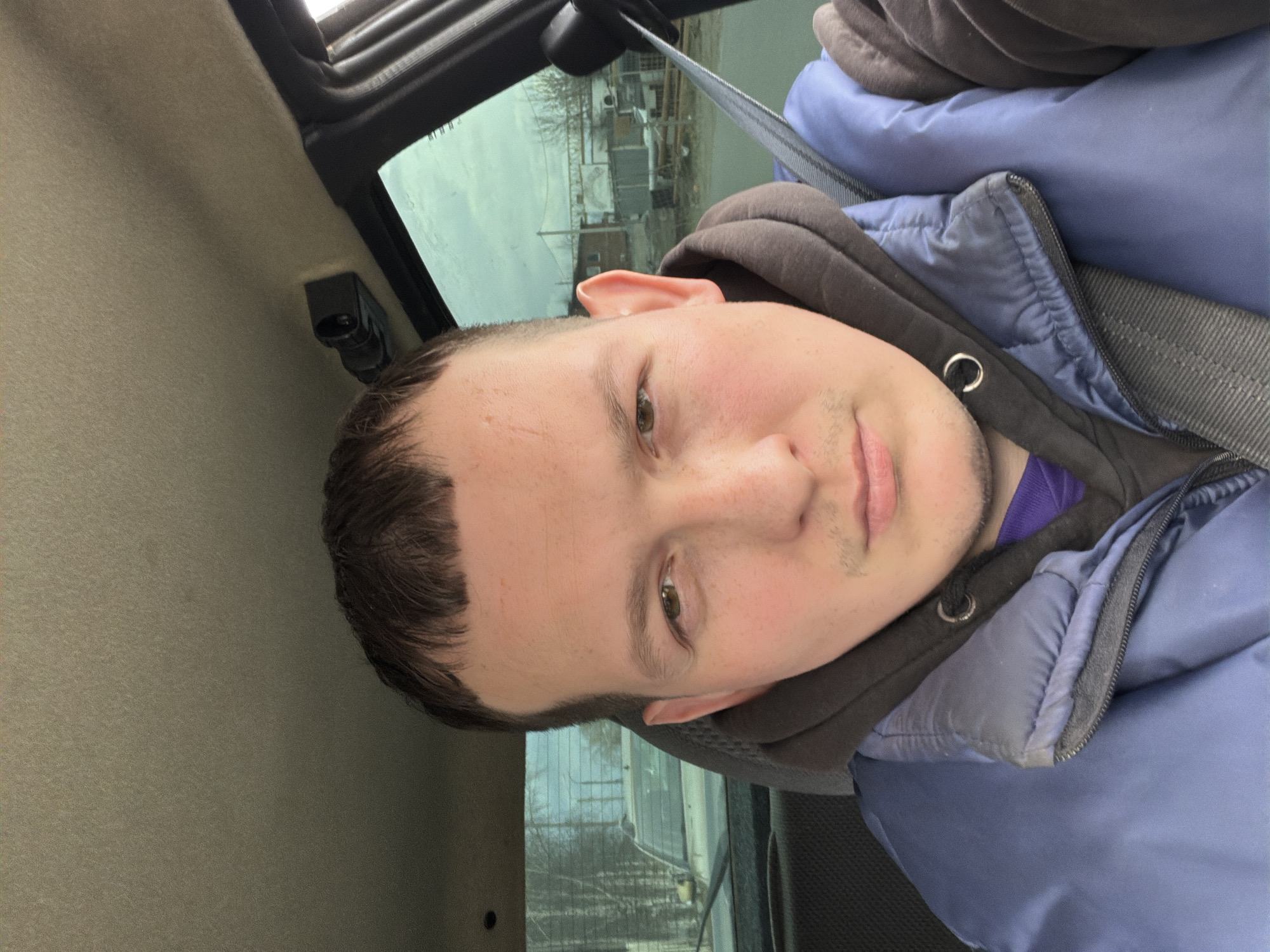 A young man is depicted in a close-up, selfie-style portrait, positioned inside a vehicle in Chaikovsky, Russia. He has light skin, short dark brown hair, and green or hazel eyes, looking directly at the camera with a neutral to slightly amiable expression. Faint stubble is visible on his chin and upper lip.
He is dressed in a dark hooded sweatshirt over a purple shirt, topped with a blue puffer jacket, suggesting cool weather. A grey seatbelt crosses his chest. The vehicle's interior features a light beige/grey headliner and a black overhead component. Through the car window, an overcast sky is visible, along with distant buildings and bare trees, reinforcing the impression of late autumn or winter. The background hints at an urban or industrial area. The activity is simply posing for the photograph. The time of day appears to be daytime, with soft, diffused lighting. There is no visible text in the image.