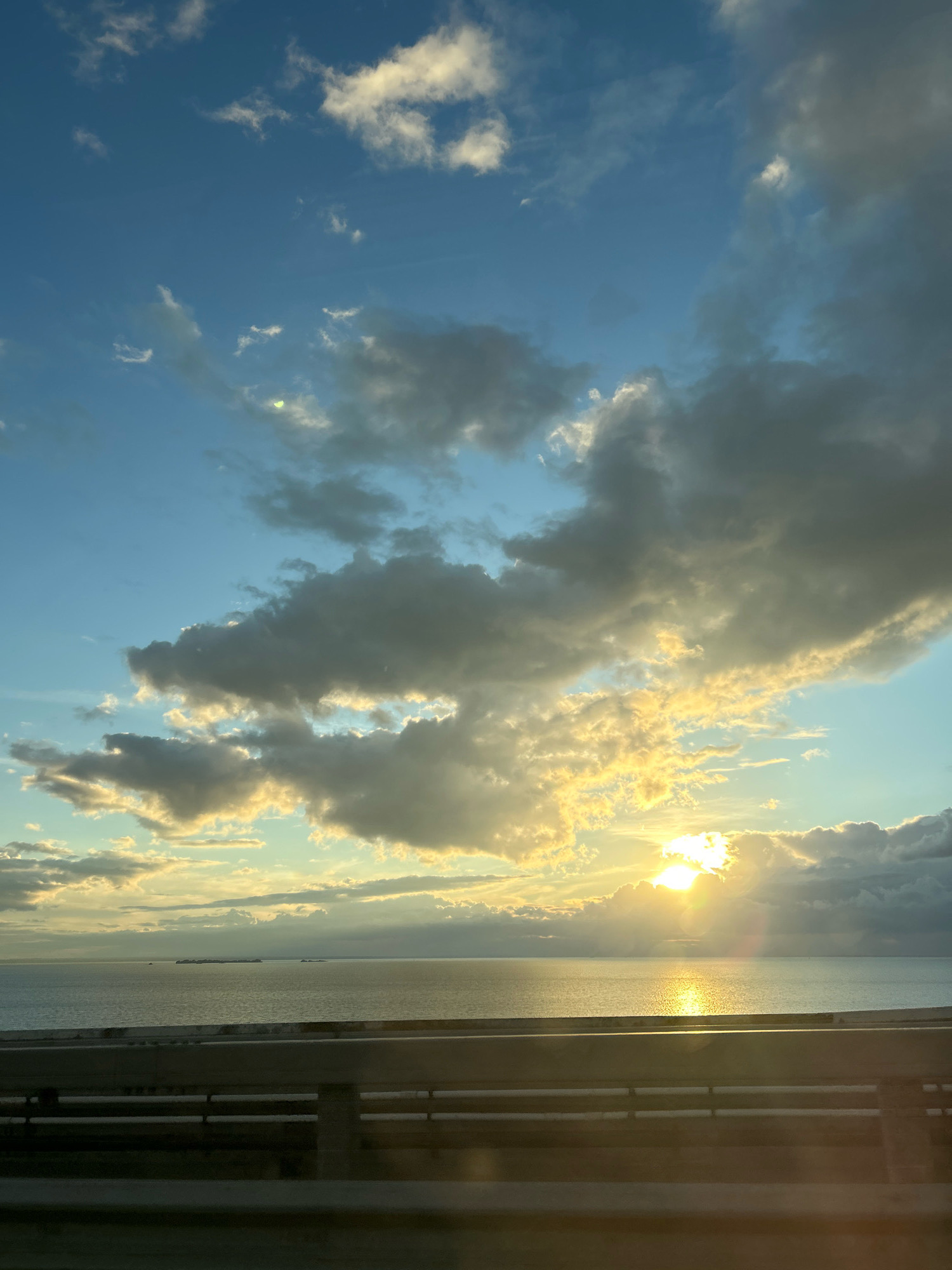 A vertical, eye-level shot captures a serene sunset over the Gulf of Finland. The sky is a vibrant gradient of blues and yellows, with a prominent sun peeking through dramatic, sunlit clouds, casting a shimmering reflection on the calm water. In the distance, a faint line of ships or islands breaks the horizon. In the foreground, a concrete barrier with horizontal railings suggests a bridge or elevated roadway, implying a location within the city of Saint Petersburg, Russia. The scene is devoid of any people or discernible activity, focusing solely on the natural beauty of the sky and sea at dusk.